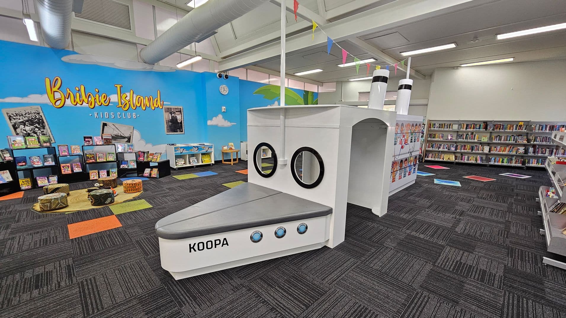 A children's library area with a nautical theme. On the left, a blue wall with "Bribie Island Kids Club" written in yellow, surrounded by bookshelves filled with colourful books. In the centre, a white play structure resembling a ship with portholes and labelled "KOOPA". The floor is carpeted in dark grey with colourful square accents. The ceiling has exposed ducts and fluorescent lighting. On the right, more bookshelves line the wall.