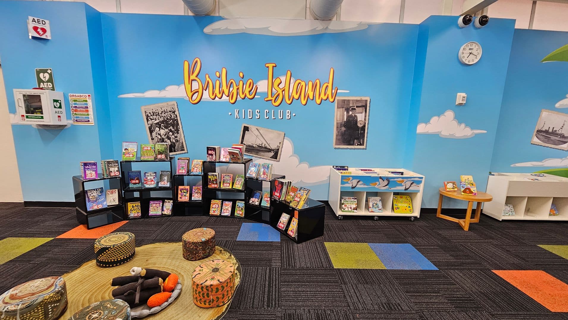 A children's reading area with a blue wall featuring the words "Bribie Island Kids Club" in large letters. Below, black shelves display various children's books. To the right, white shelves and a small round table hold more books. The floor is covered with a colourful carpet in shades of purple, green, blue, and orange. Cushioned stools with patterns are arranged around a circular mat. A clock and AED signs are mounted on the wall. Vintage photos are also displayed on the wall.
