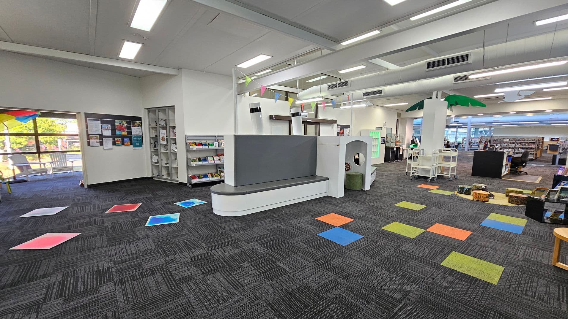 A library interior with a grey carpeted floor featuring colourful square tiles. On the left, a window with a view outside and a notice board with various papers. A bookshelf with books and magazines is nearby. In the centre, a white and grey seating area with a play structure. On the right, a palm tree decoration, book carts, and baskets on the floor. The ceiling has fluorescent lights and exposed ducts.