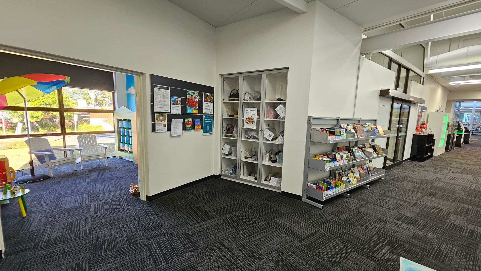 On the left, a room with large windows, white chairs, and a colourful umbrella. A small table with toys is visible. In the centre, a wall with a bulletin board displaying various notices and a shelf with toy collections. On the right, a bookshelf filled with books and a glass door leading to another area. The floor is covered with dark patterned carpet tiles, and the ceiling has exposed beams and lighting fixtures.