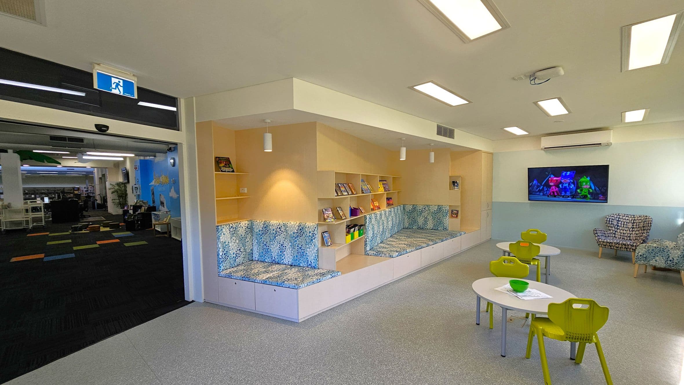 A children's reading area with a light grey floor and a ceiling with rectangular lights. On the left, there is an open entrance with a blue exit sign above, leading to a room with colourful carpet tiles. In the centre, there are built-in bookshelves with books and cushioned seating with a blue pattern. On the right, a wall-mounted TV displays an animated show above two small tables with green chairs. A patterned armchair is nearby.