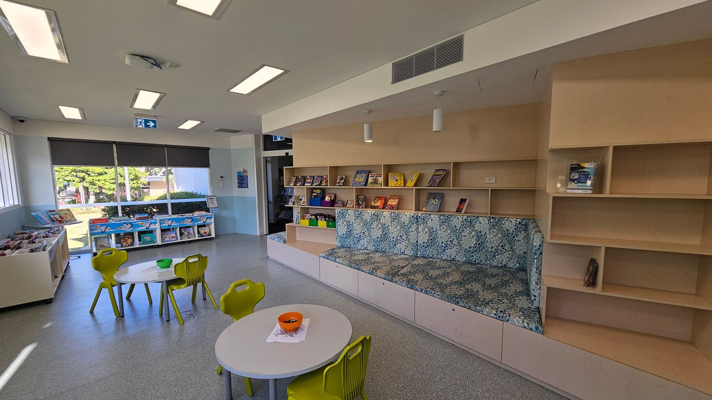 On the left, there are large windows with a view of trees and a building outside. Below the windows, there are shelves filled with books. In the centre, two small round tables with green chairs are placed on a grey floor, with a bowl on one table. On the right, there is a built-in wooden bookshelf with books and a cushioned seating area with a patterned fabric. The ceiling has rectangular lights and an air vent.