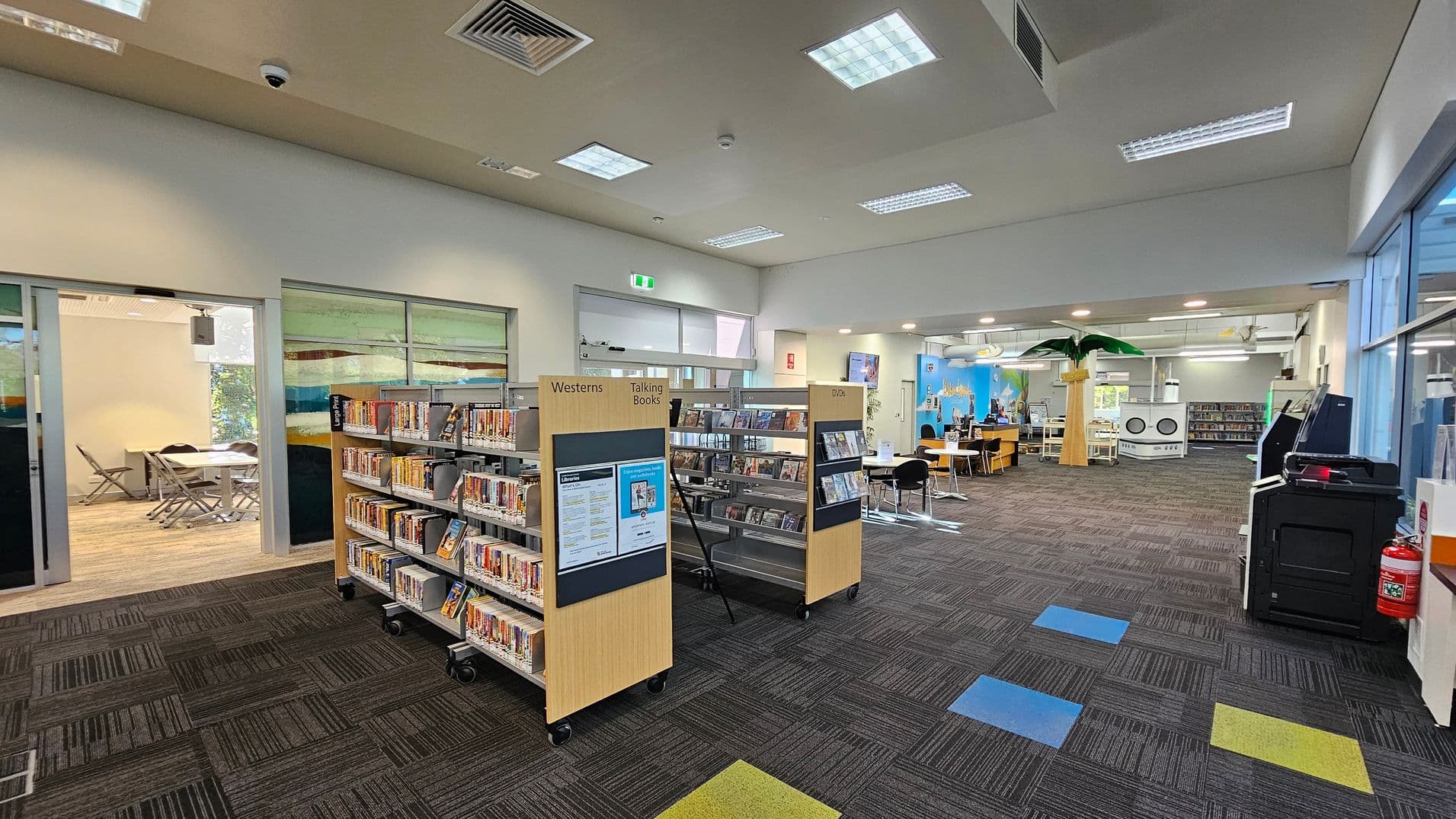 A library interior with a patterned carpet floor featuring blue and yellow squares. On the left, there are bookshelves labelled "Westerns" and "Talking Books," with DVDs on the right. In the background, a room with tables and chairs is visible through glass doors. The right side has a large palm tree decoration and a seating area with tables and chairs. The ceiling has recessed lighting and air vents. A fire extinguisher and a black machine are on the far right.