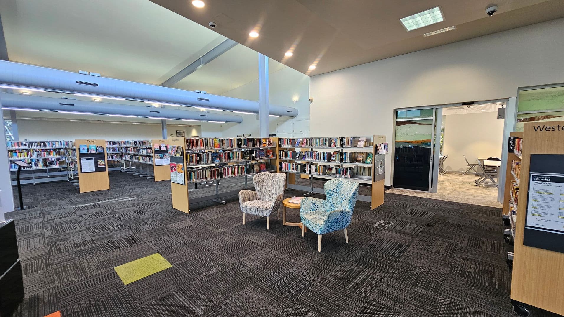 A library interior with bookshelves filled with books on the left and right. In the centre, two patterned armchairs are positioned around a small round table. The floor is covered with dark, patterned carpet tiles. In the background, a glass-walled room with tables and chairs is visible. The ceiling is high with recessed lighting and large, exposed ducts.