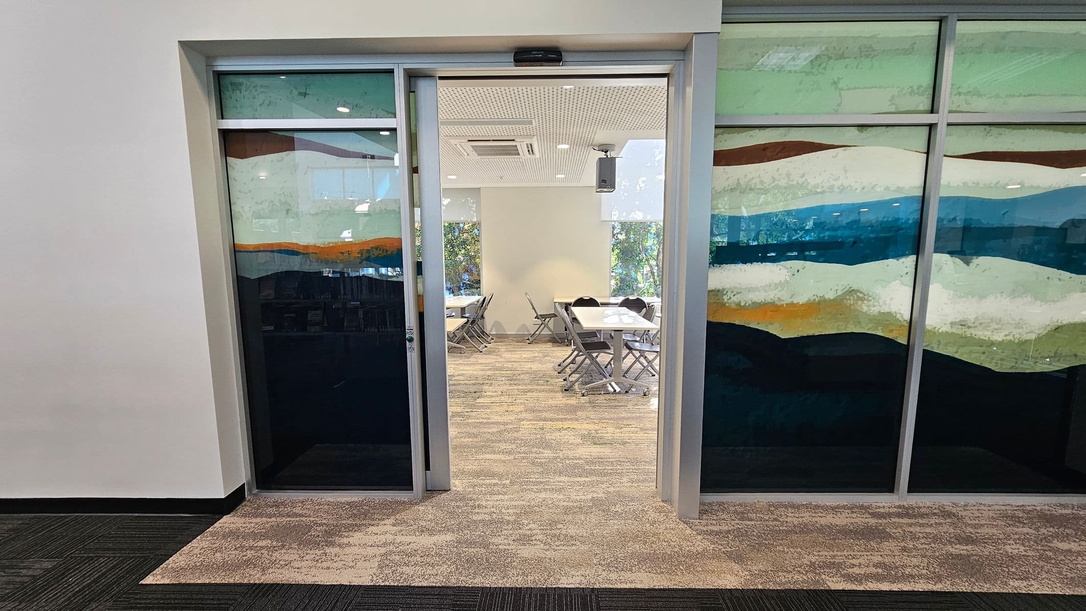 A room with glass sliding doors featuring a colourful abstract design. Inside, there are several tables and chairs arranged in a meeting or classroom style. The floor is covered with a patterned carpet, and the ceiling has recessed lighting and an air conditioning unit. The walls are light-coloured, and there are windows in the background allowing natural light to enter.