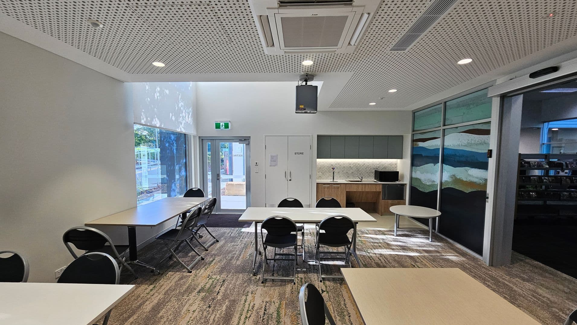 A room with a perforated ceiling featuring recessed lights and an air conditioning unit. On the left, large windows allow natural light in, illuminating tables and chairs. In the centre, a door marked "Store" is flanked by a kitchenette with cabinets, a sink, and a microwave. On the right, a wall with abstract art panels and a round table. The floor is carpeted with a patterned design.