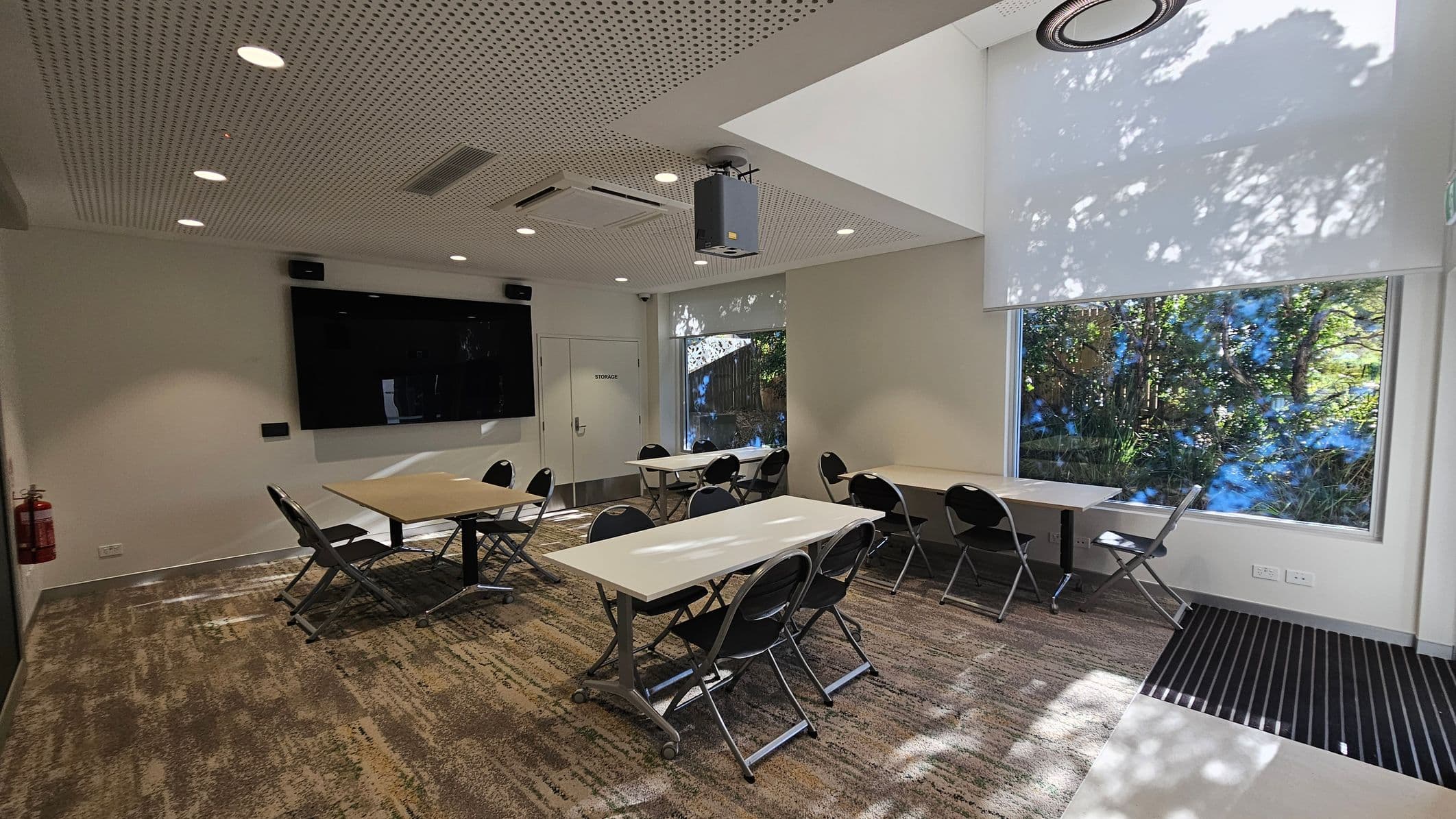 A modern conference room with a perforated ceiling featuring recessed lights and a projector. On the left, a large black screen is mounted on the wall above a door labelled "Storage". Several folding tables and chairs are arranged in the centre. On the right, large windows with roller blinds partially down allow natural light to filter through, casting shadows of trees outside. The floor is carpeted with a patterned design, and a fire extinguisher is mounted on the wall.