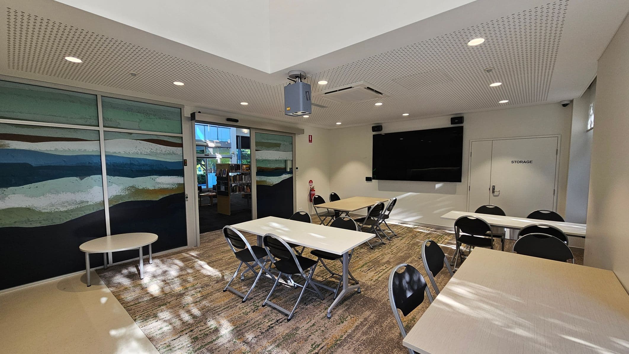 A room with a patterned carpet and a perforated ceiling with recessed lights. On the left, there is a large mural with abstract horizontal stripes in various colours. In the centre, several tables and chairs are arranged, with a projector mounted on the ceiling. On the right, a large black screen is mounted on the wall above a door labelled "STORAGE". A fire extinguisher is positioned near the door. In the background, a glass door leads to another area with bookshelves.