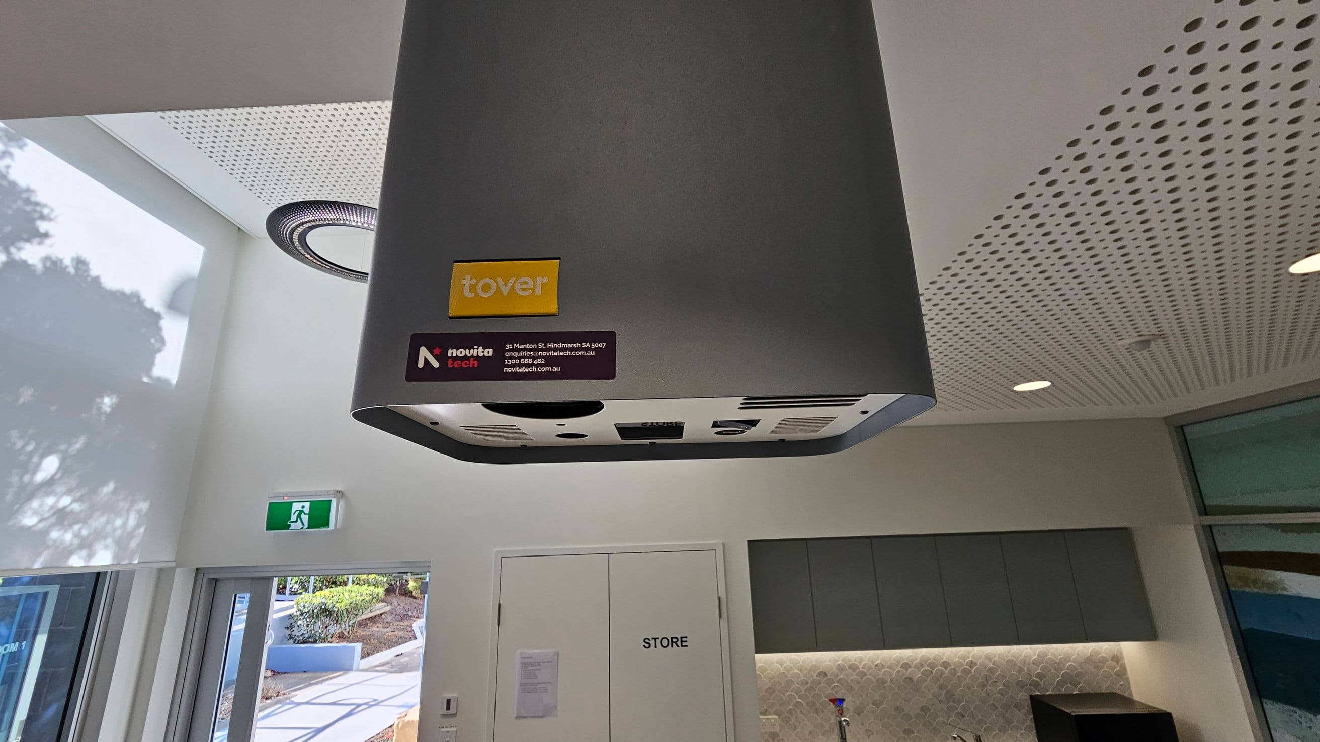 A grey ceiling-mounted device with "tover" and "novita tech" labels is in the foreground. The ceiling has a perforated design with recessed lighting. Below, a white door marked "STORE" is flanked by grey cabinets and a countertop with a sink. On the left, a glass door leads outside, where a path and greenery are visible. An emergency exit sign is above the door.
