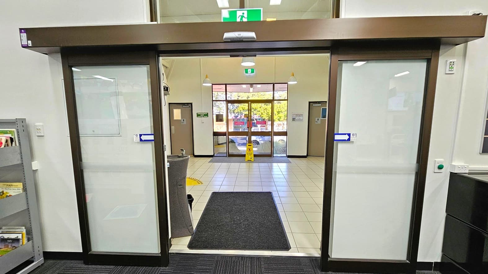 Automatic sliding glass doors with a dark frame are in the foreground, leading to a tiled entrance area. A black mat is on the floor just inside the doors. Beyond, a larger set of glass doors opens to the outside, with a yellow caution sign on the floor. On the left, a grey shelving unit holds books, and on the right, a wall with a green button and power outlet. The ceiling has recessed lighting and a green emergency exit sign above the doors.