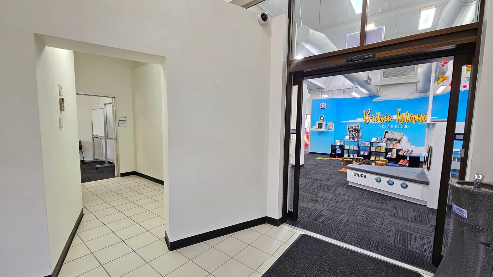 A hallway with white tiled flooring and white walls leads to a room on the left with a whiteboard visible inside. On the right, a doorway opens into a room with dark carpeted flooring. The back wall is bright blue with the words "Bribie Island Kids Club" in yellow. Below the sign, there are shelves displaying books. A play kitchen labelled "Koopa" is in the foreground. The ceiling has exposed ducts and lights, and a water fountain is visible on the right.