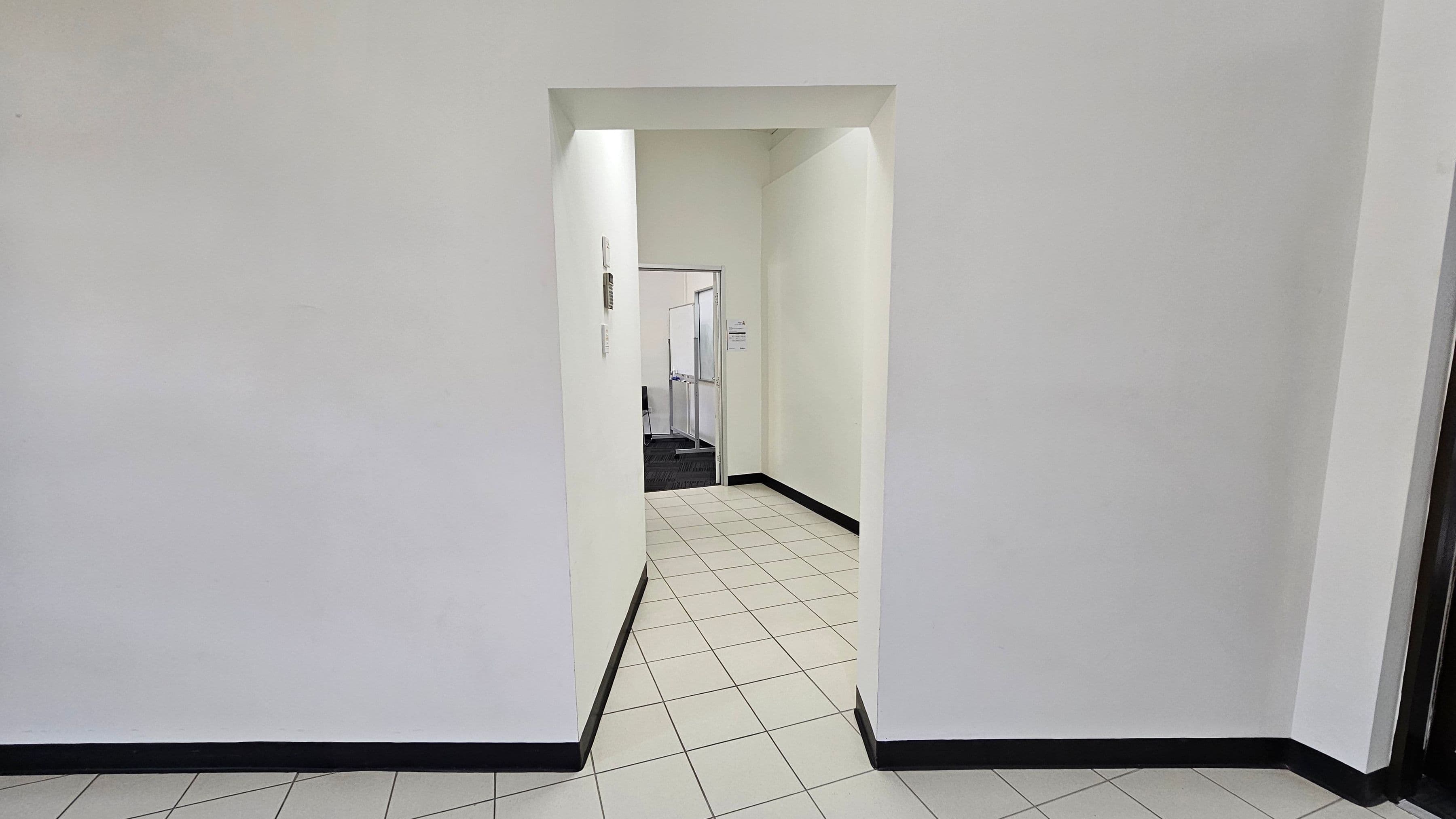 A hallway with white walls and a tiled floor leading to an open doorway. Beyond the doorway, there is a room with a darker carpeted floor. The walls have a slight yellow tint, and there are some fixtures on the left wall near the doorway. The ceiling is plain and white, with no visible features.