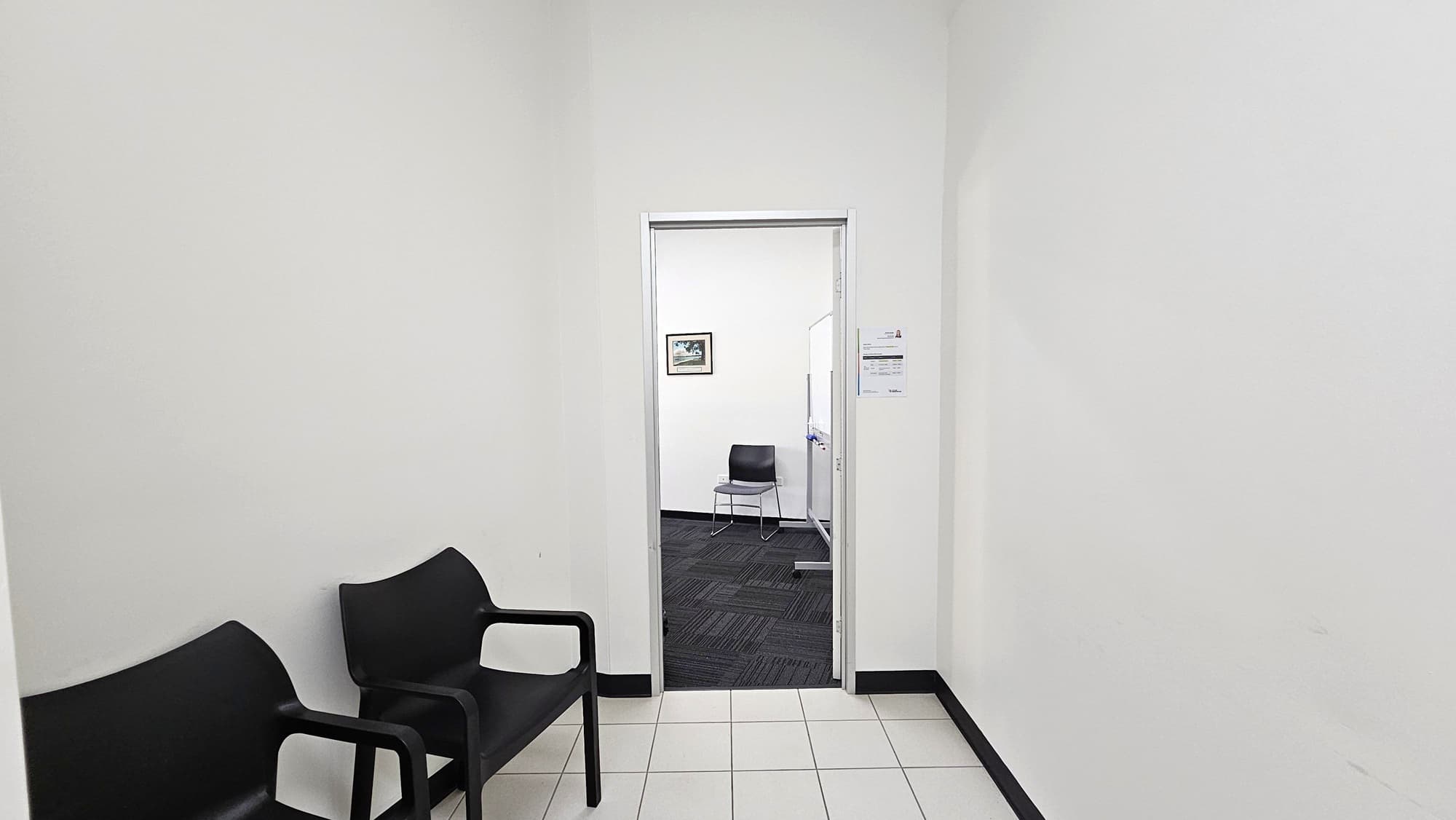 A hallway with white walls and a tiled floor. On the left, there are two black chairs. Ahead, an open doorway leads to another room with a dark carpeted floor. Inside, a single black chair is visible against the far wall. A framed picture hangs on the wall to the left, and a whiteboard is partially visible on the right. A small sign is posted on the wall near the doorway.