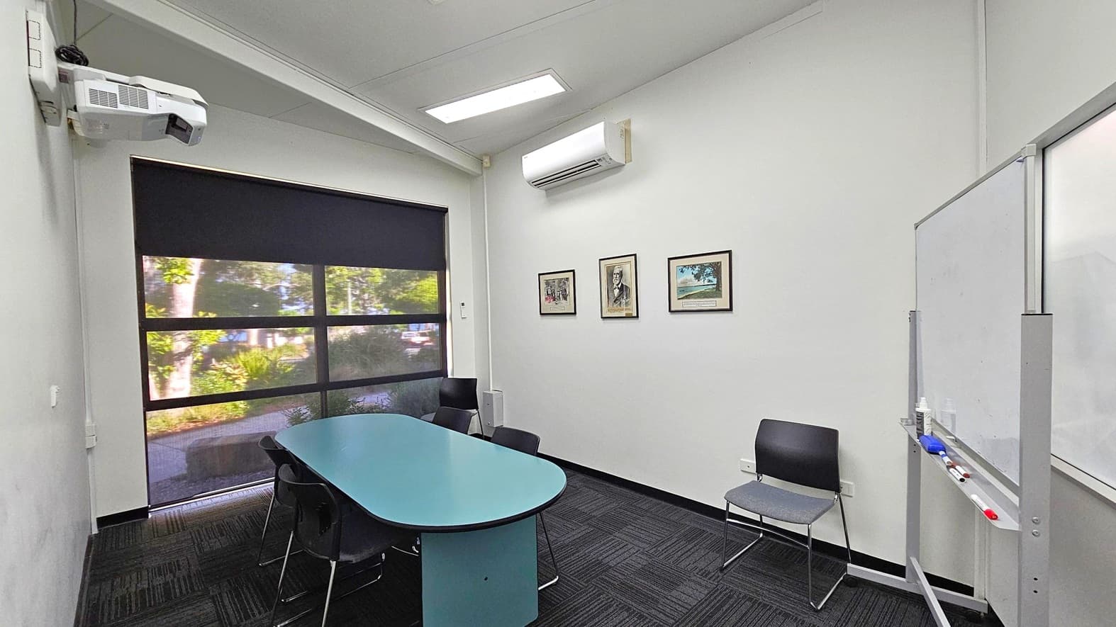 A small meeting room with a black carpeted floor and a white ceiling. On the left, a window with a black roller blind partially open, revealing greenery outside. Below the window is a blue oval table surrounded by black chairs. On the right wall, three framed pictures are displayed. An air conditioning unit is mounted above them. A whiteboard with markers is positioned on the far right. A projector is mounted on the ceiling.
