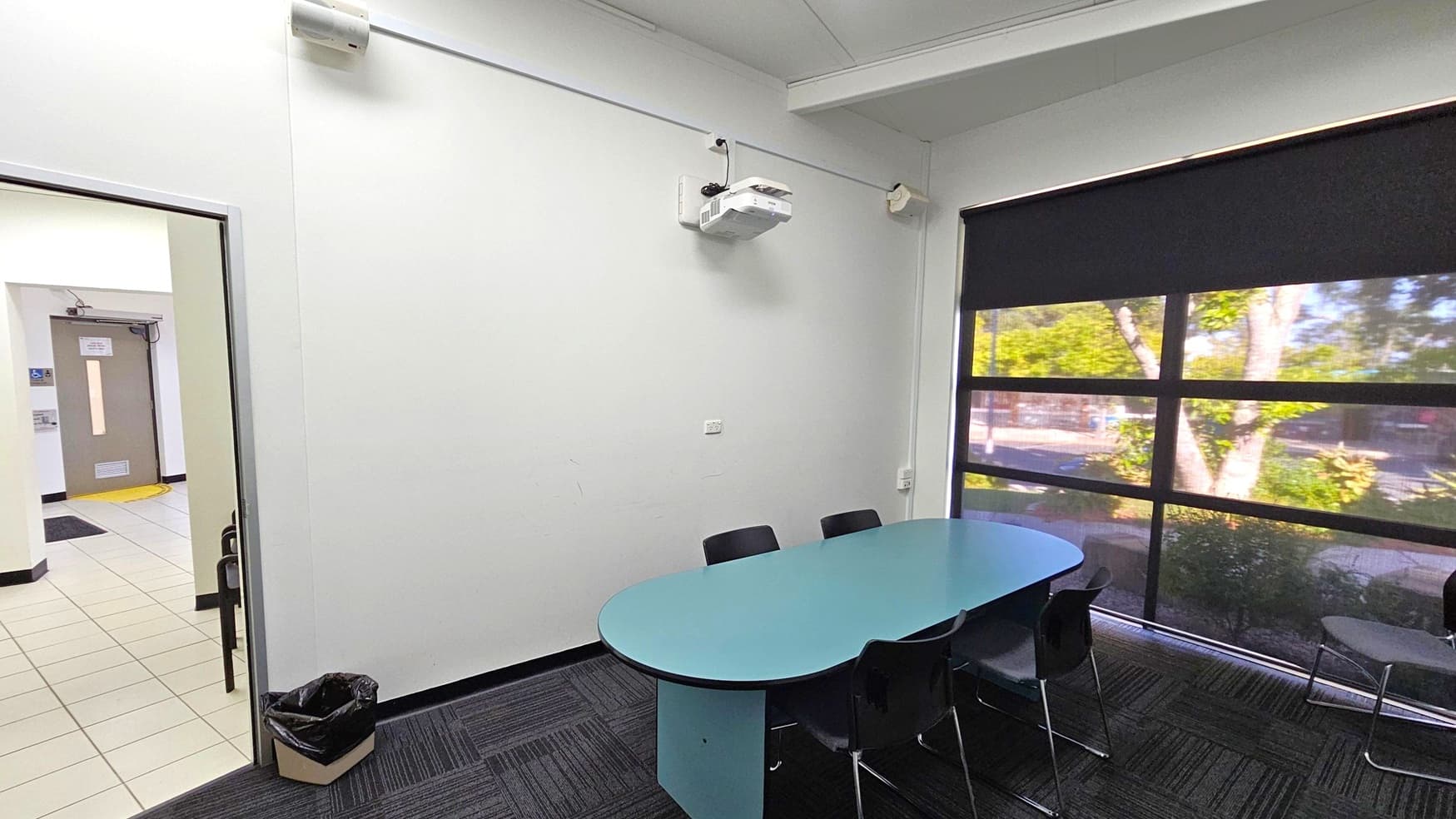 A small meeting room with a blue oval table surrounded by black chairs on the right. A large window with a black blind partially drawn reveals trees outside. On the left, an open door leads to a tiled hallway with a brown door visible. A black bin is on the floor near the door. A white projector is mounted on the wall above the table. The floor is carpeted with a dark pattern, and the ceiling is white with visible beams.