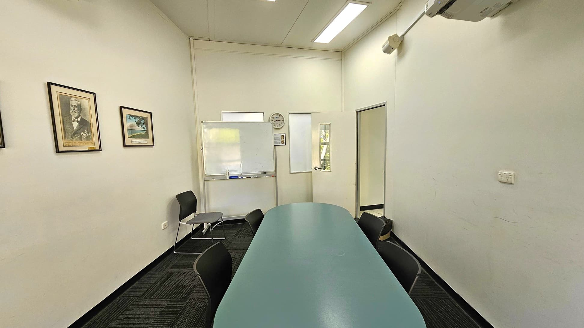 A small meeting room with a long oval table in the centre surrounded by black chairs. On the left wall, there are three framed pictures. The back wall features a whiteboard, a clock, and a door with a window. The right wall has an air conditioning unit mounted near the ceiling and a power outlet. The floor is carpeted in a dark pattern, and the ceiling has recessed lighting.