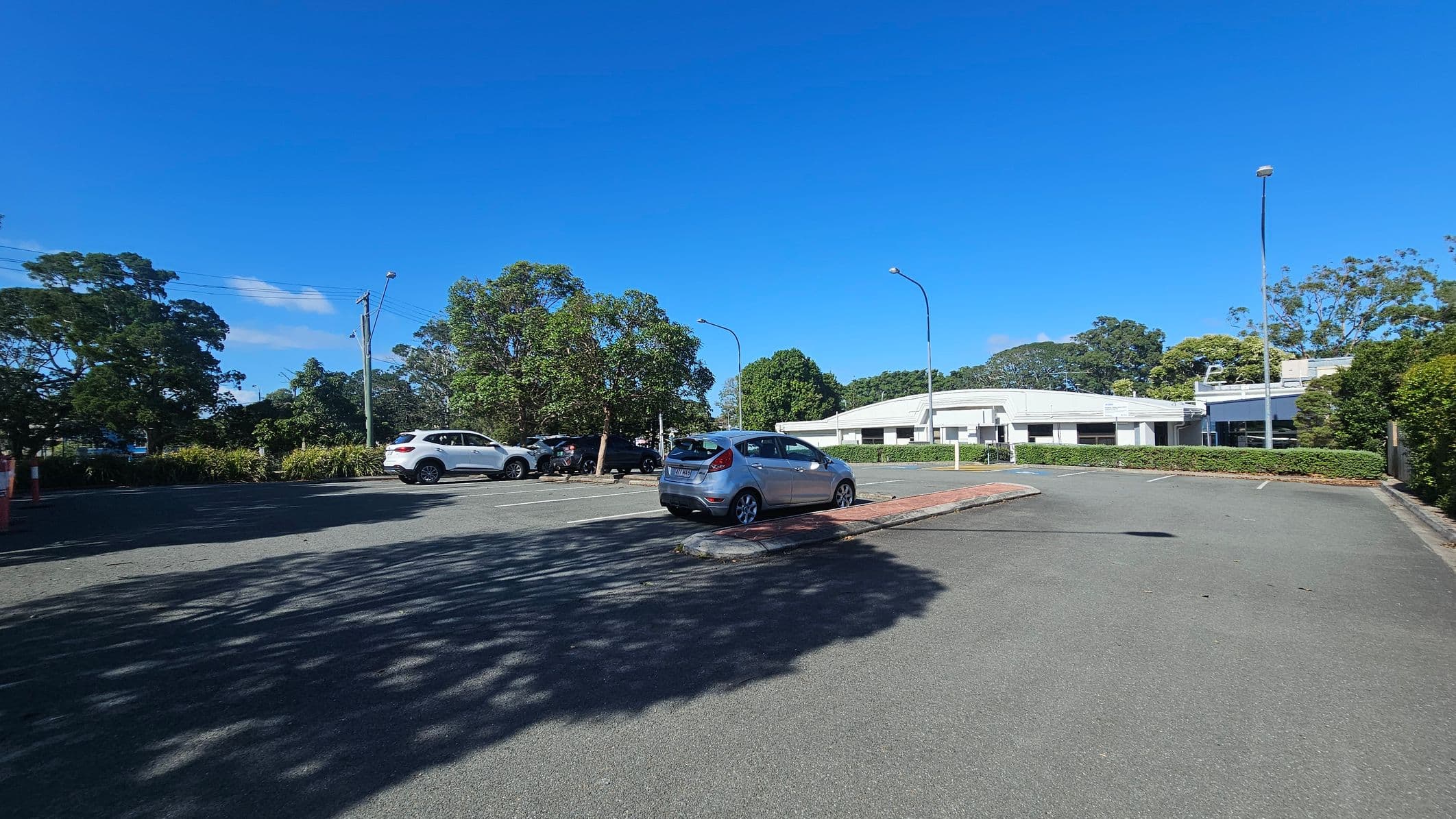 A car park with a few vehicles, including a silver car in the centre. On the left, there are trees and a white SUV. In the background, a white building is partially visible, surrounded by more trees and greenery. The ground is paved, and there are streetlights along the perimeter. Shadows of trees are cast on the pavement.