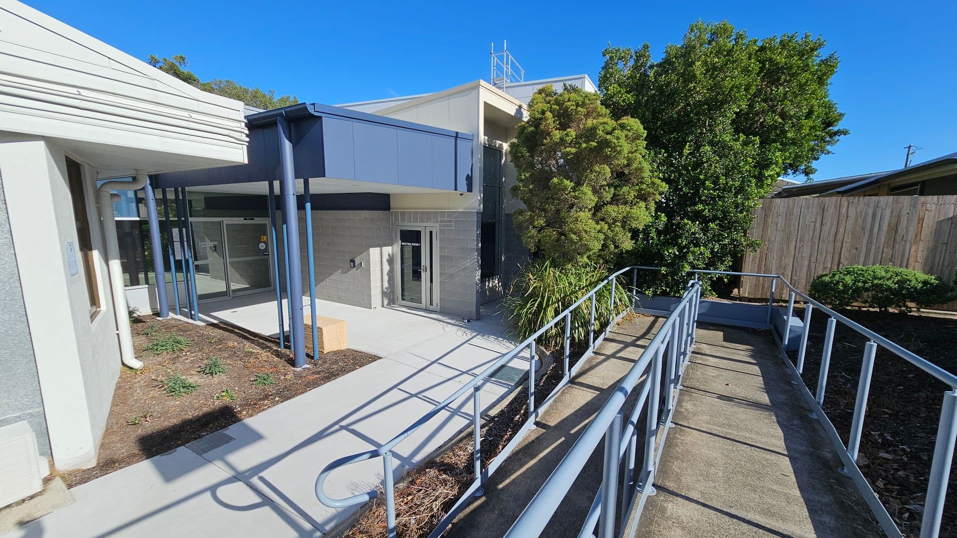 A building entrance with a ramp leading up to it. On the left, there is a white and grey wall with a window and a downpipe. The middle features a glass door with a sign, supported by blue pillars and a small garden area with plants. On the right, there is a wooden fence and dense greenery, including a large bush and smaller shrubs. The ramp has metal railings and is bordered by a narrow garden bed. The ground is paved with concrete.