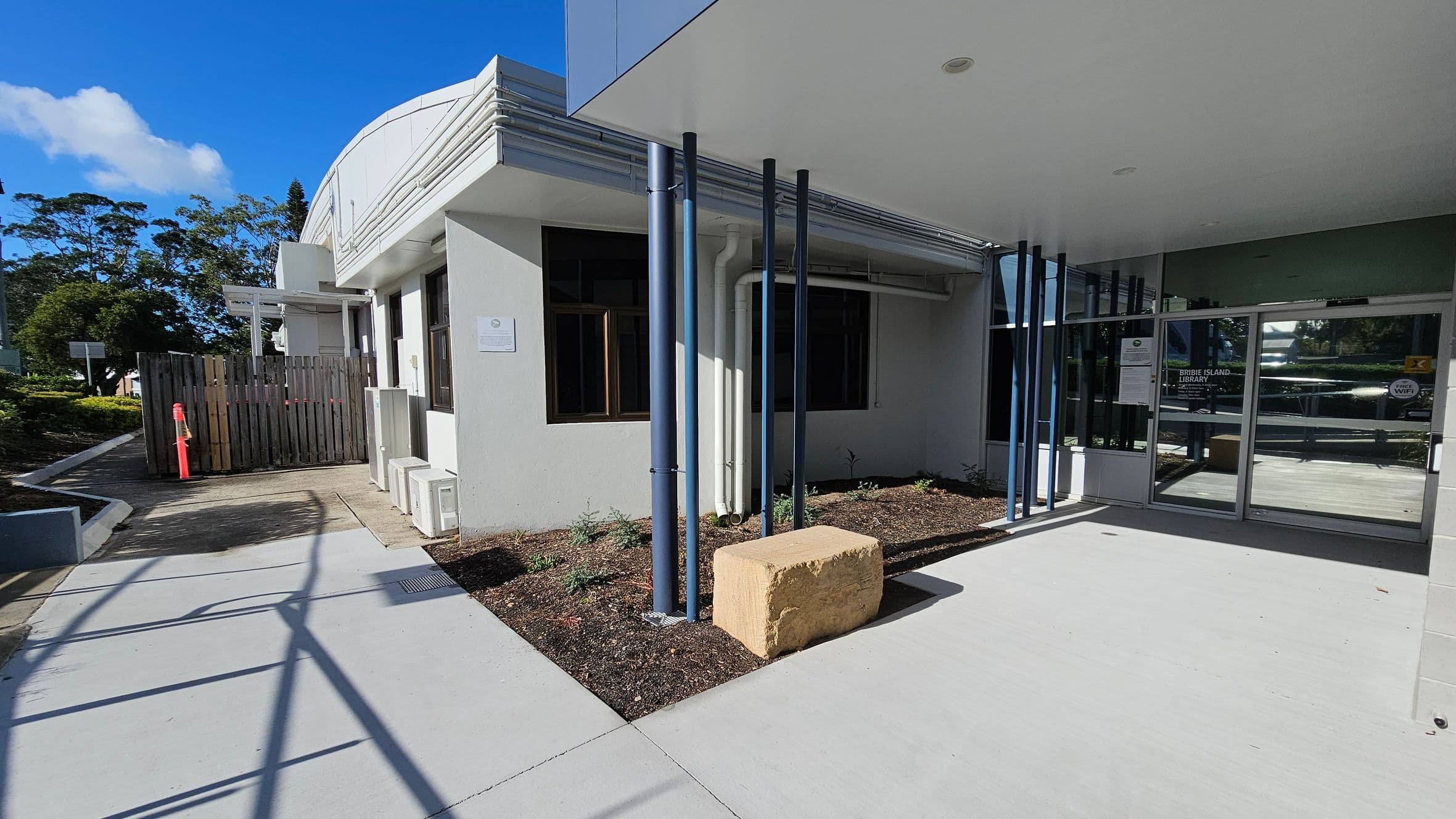 A modern building entrance with a glass door on the right, featuring signage. Blue vertical pipes are positioned near the entrance, with a large rectangular stone block beside them. The left side has a wooden fence and a pathway leading away from the building. The ground is concrete, and there are small plants in a mulched area near the building. The ceiling is flat and white, with a recessed light. Trees and greenery are visible in the background.