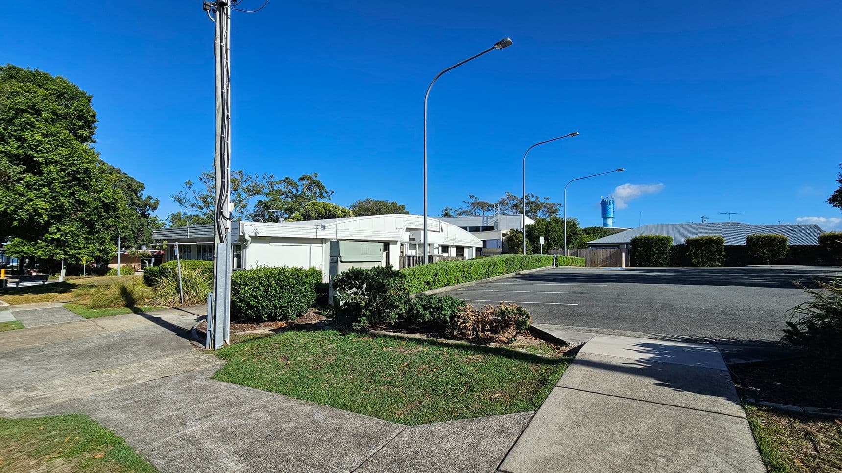 A street scene with a white building in the centre, surrounded by green hedges and trees. On the left, a tall utility pole stands near a grassy area and a concrete footpath. The right side features a paved parking area with several streetlights. In the background, a blue structure is visible, partially obscured by trees and buildings. The sky is clear and blue.