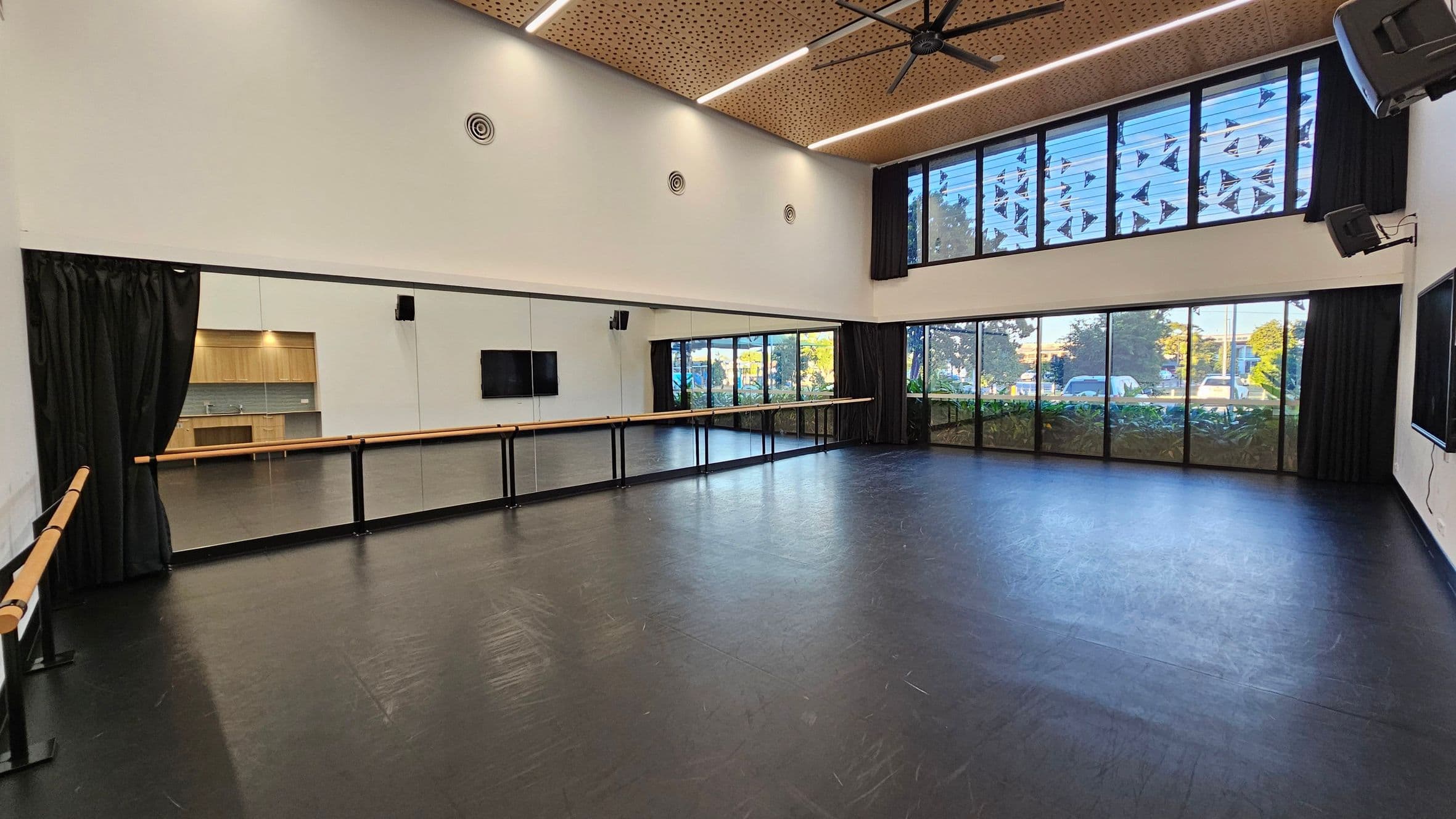 A spacious dance studio with a black floor and a high ceiling featuring wooden panels and a large fan. The left wall has a long mirror with a wooden ballet barre running across it. The right side has large windows with black curtains, allowing natural light to enter. In the background, there is a kitchenette with wooden cabinets and a small sink. The studio is equipped with speakers and a wall-mounted screen.