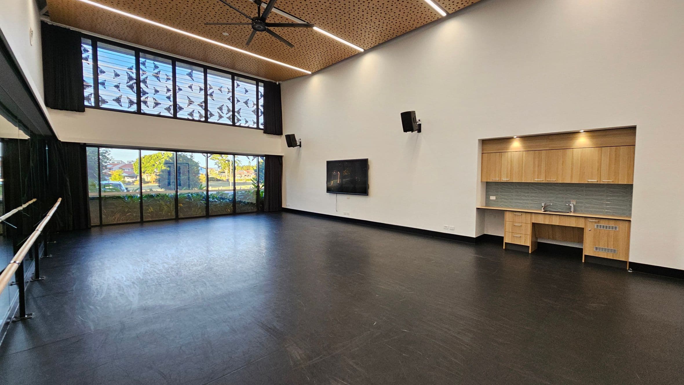A spacious room with a dark floor and a high ceiling featuring wooden panels and recessed lighting. On the left, there are large windows with patterned designs and a ballet barre. The back wall has floor-to-ceiling windows with a view of trees and a building. On the right, there's a wall-mounted screen and a kitchenette with wooden cabinets, a sink, and a tiled backsplash. Ceiling fans are visible above.