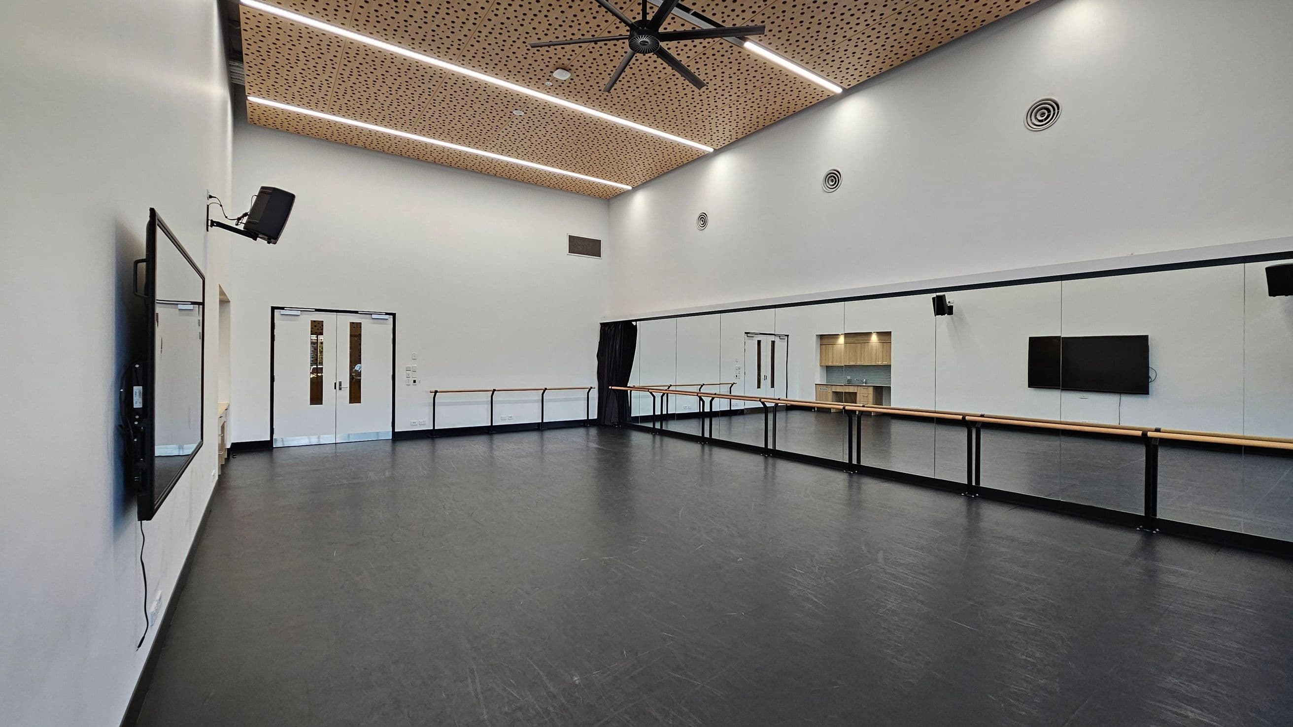 A spacious dance studio with a black floor and white walls. On the left, a large wall-mounted screen and a speaker are visible. The right side features a long mirror with a wooden ballet barre running along it. In the centre, double doors with narrow windows are present. The ceiling is wooden with a perforated design, featuring linear lights and a large ceiling fan.