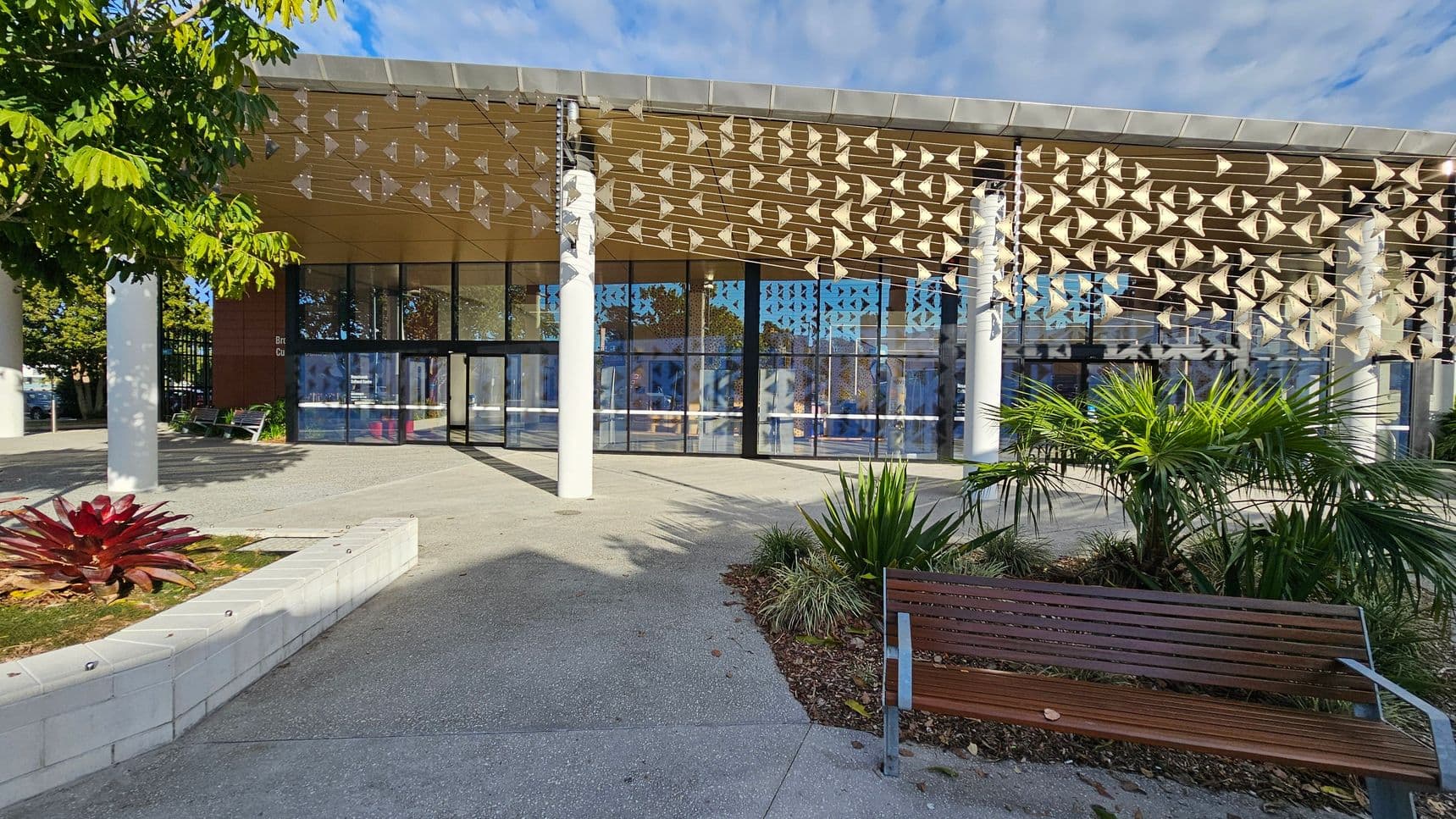 A modern building with a glass facade and a decorative overhang featuring geometric patterns. In the foreground, there is a wooden bench on the right and a landscaped area with various plants, including a red-leaved plant on the left. The ground is paved with concrete, and there are white columns supporting the structure. A tree with green leaves is partially visible on the left side.
