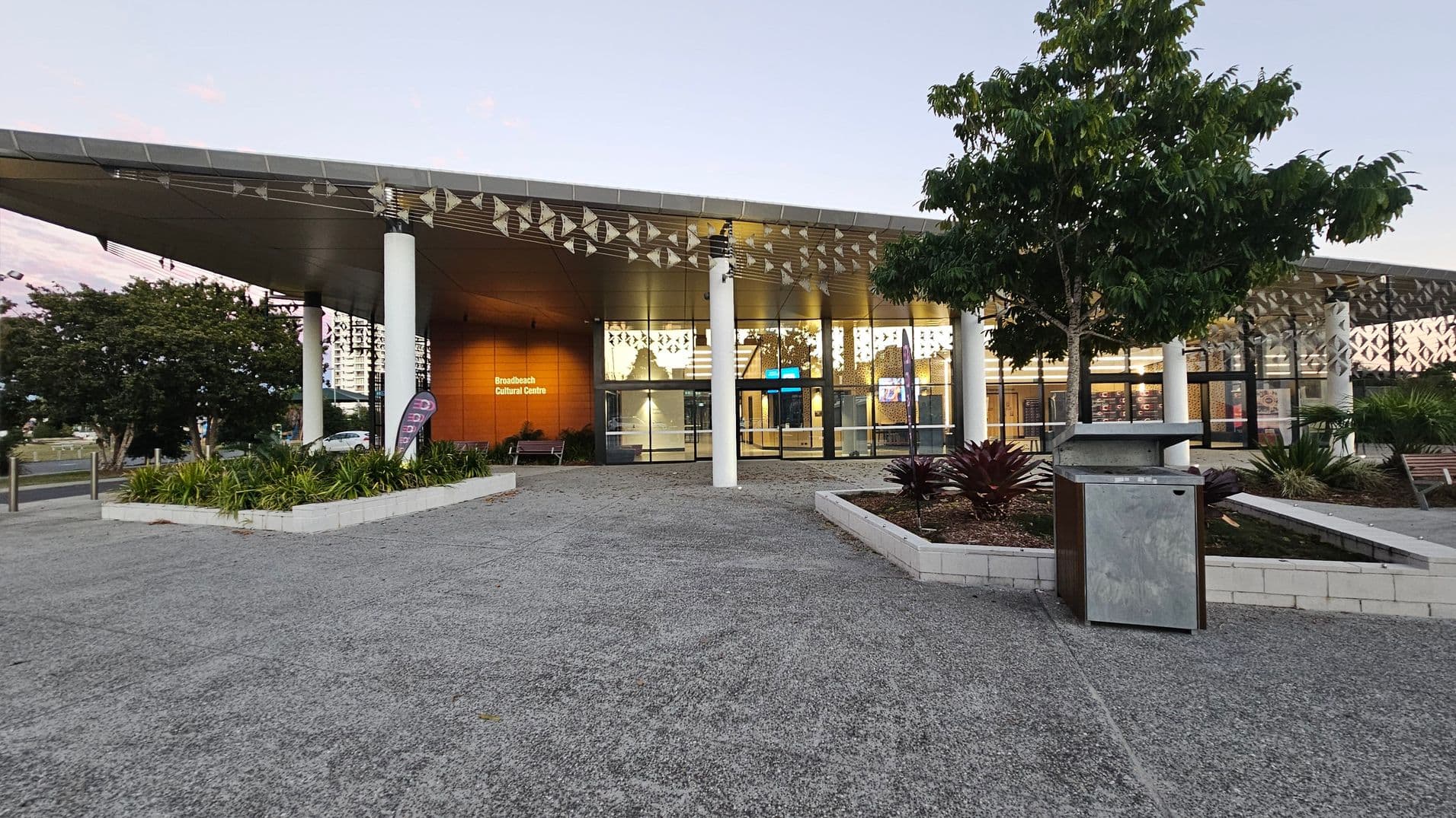 A modern building with a wide, overhanging roof supported by white columns. The entrance features large glass windows and doors, with a sign reading "Broadbeach Cultural Centre" on a wooden panel. In the foreground, there are landscaped garden beds with green plants and a tree. A concrete path leads to the entrance, and a metal bin is positioned near the garden. The ceiling has a decorative pattern, and the area is well-lit from inside.