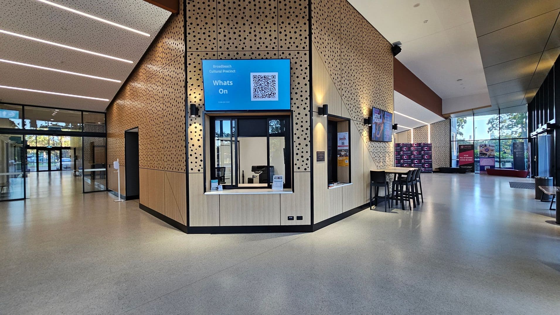 A modern interior with a perforated wood-panelled wall featuring two service windows. Above one window is a blue sign with a QR code. The floor is smooth and light-coloured. On the right, there's a high table with stools and a wall-mounted screen. Large glass windows in the background allow natural light, with banners and a bench visible outside. The ceiling is angled with linear lighting.