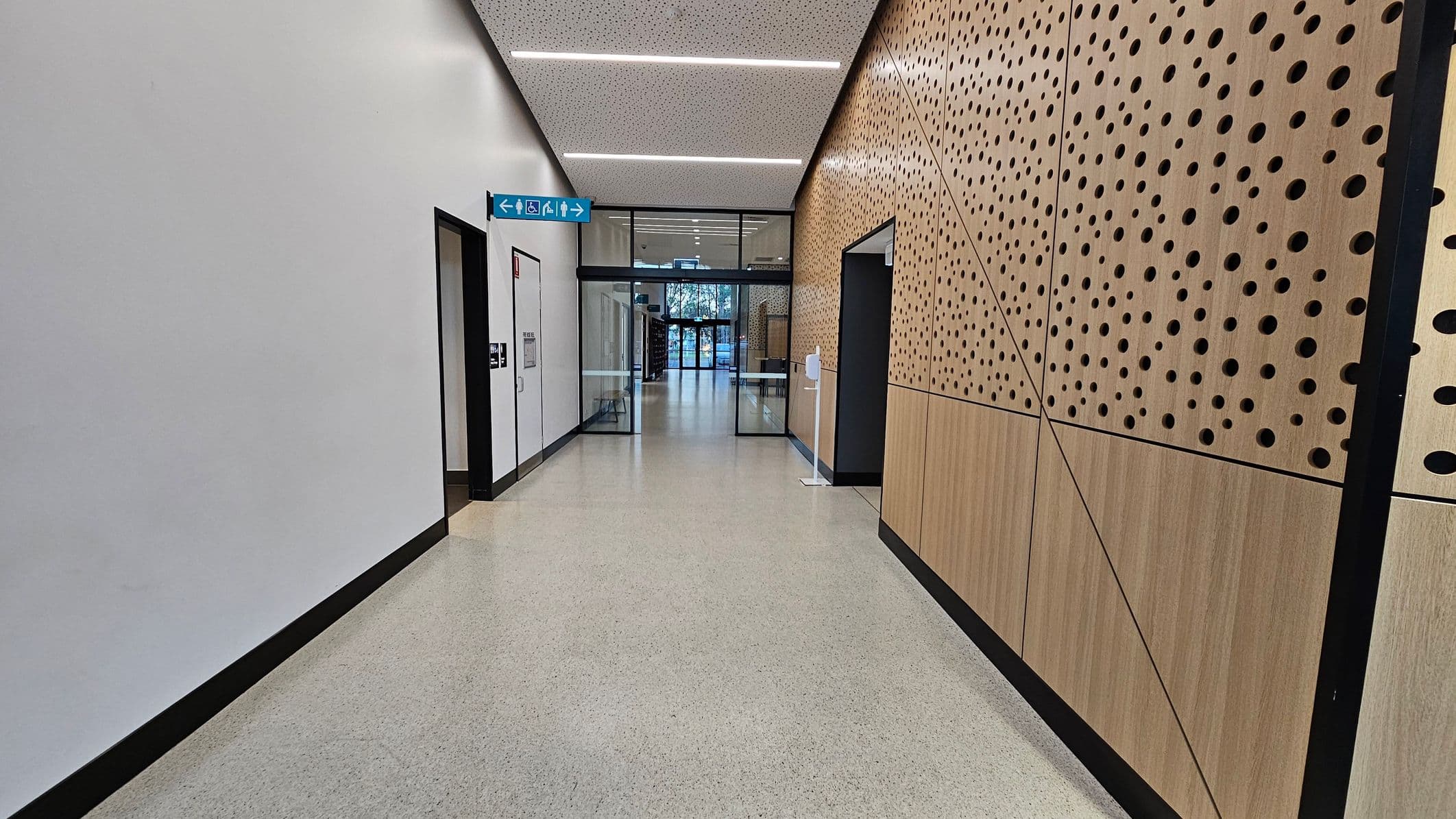 A hallway with a light-coloured floor and a perforated wooden panel wall on the right. The left wall is plain white with a door. Overhead, a ceiling with linear lights. At the end of the hallway, glass doors lead to an area with visible trees outside. A blue sign with directional symbols is mounted near the ceiling. A hand sanitiser dispenser is attached to the right wall.