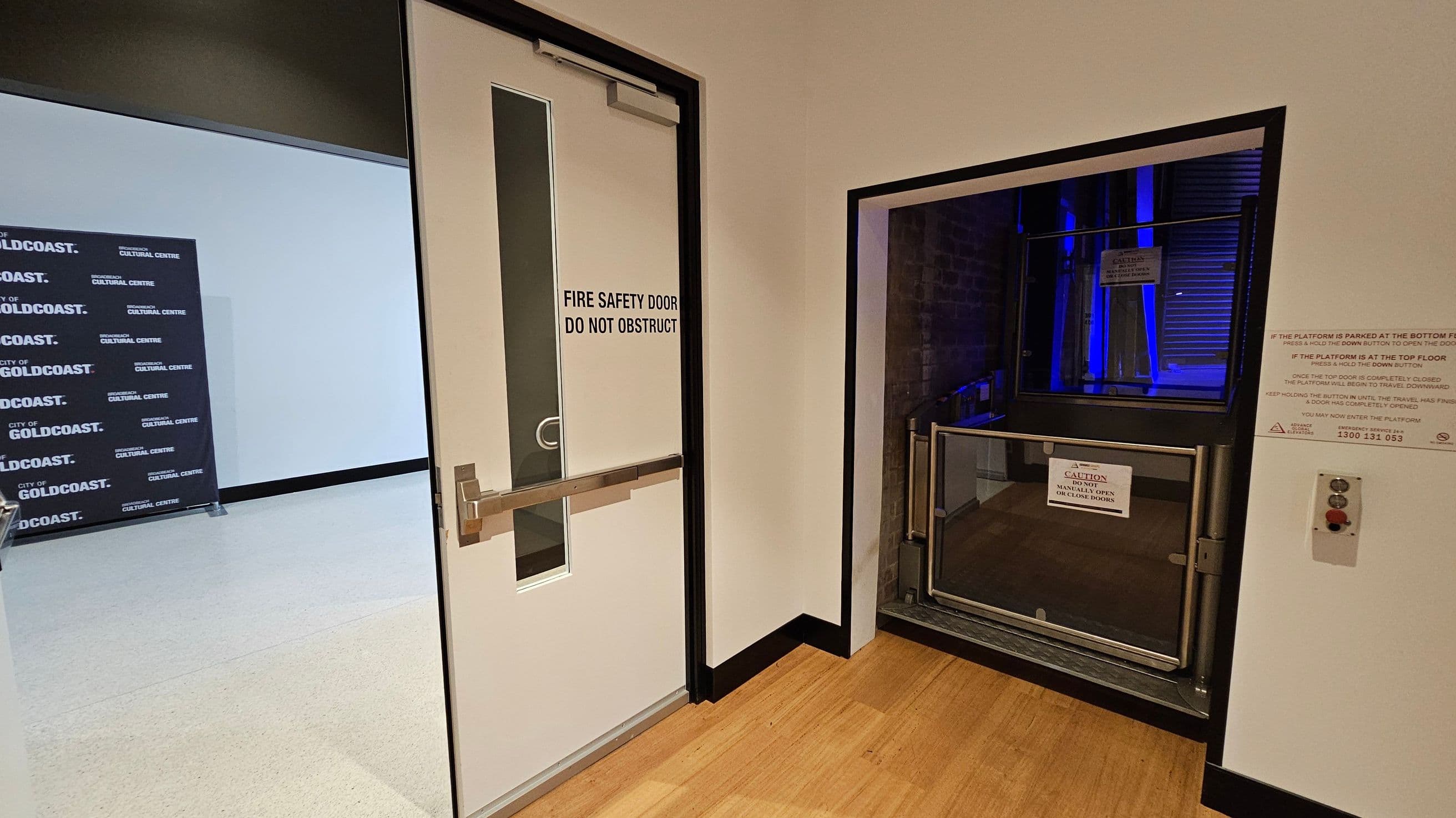 A white fire safety door with a vertical window and a push bar is on the left, marked "DO NOT OBSTRUCT." To the right, there's a platform lift with a caution sign, surrounded by metal railings. The lift area is illuminated with blue lighting. A control panel with buttons is mounted on the wall next to the lift. The floor is wooden, and the walls are light-coloured. In the background, a sign reads "CITY OF GOLDCOAST CULTURAL CENTRE."
