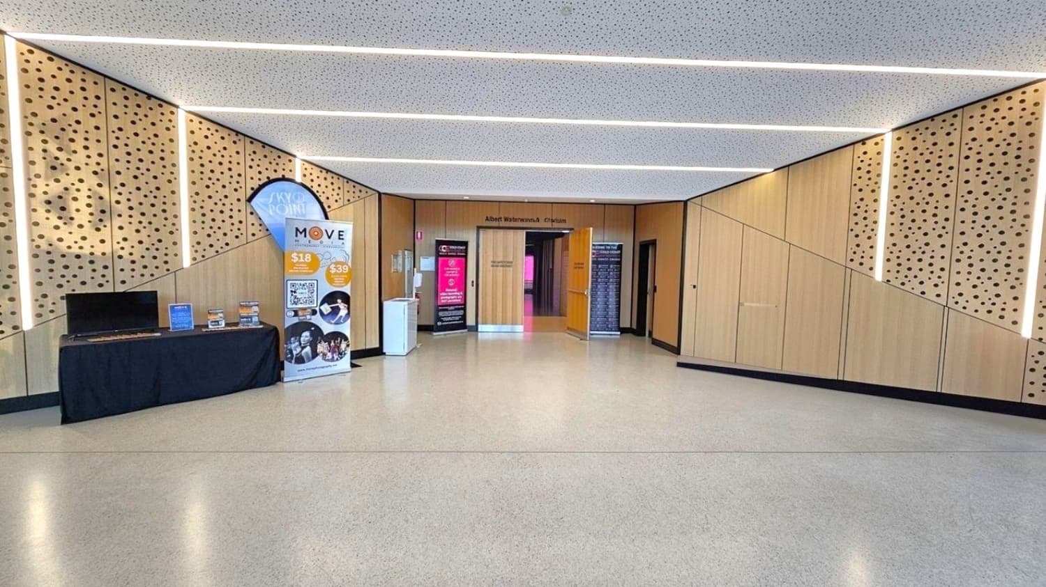 A hallway with a light-coloured, speckled floor and a perforated ceiling. The walls are wooden with circular patterns. On the left, a table with a black cloth holds promotional materials, and a banner stands nearby. In the centre, double doors lead to the Albert Waterways Auditorium, flanked by more banners.