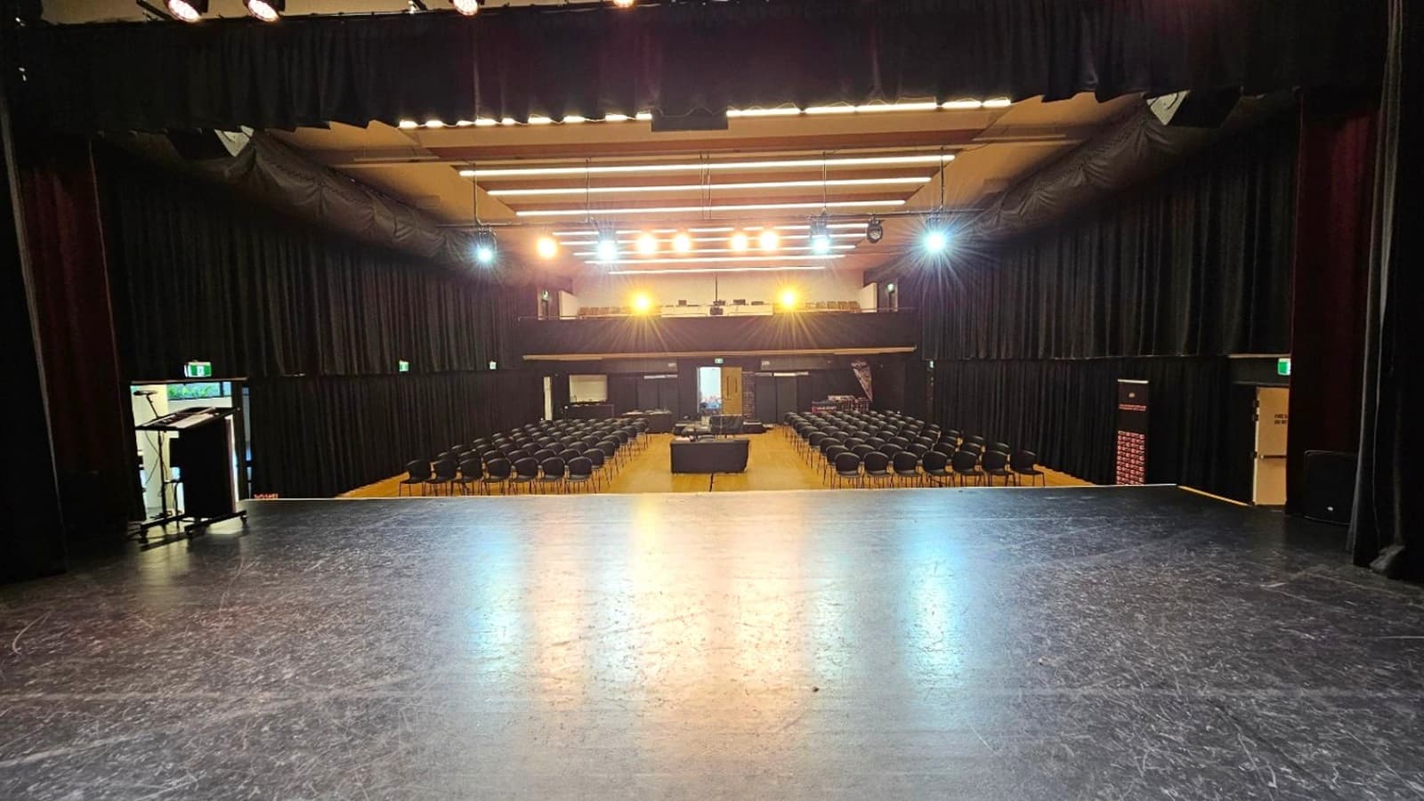 View from a stage looking out into an auditorium. The foreground features a dark, polished stage floor. In the middle, rows of empty black chairs are arranged facing the stage, with a central aisle. The background shows a balcony with additional seating and bright stage lights illuminating the space. The walls and ceiling are lined with black curtains, and there are exit signs visible on the sides. A podium is positioned to the left near an exit door.