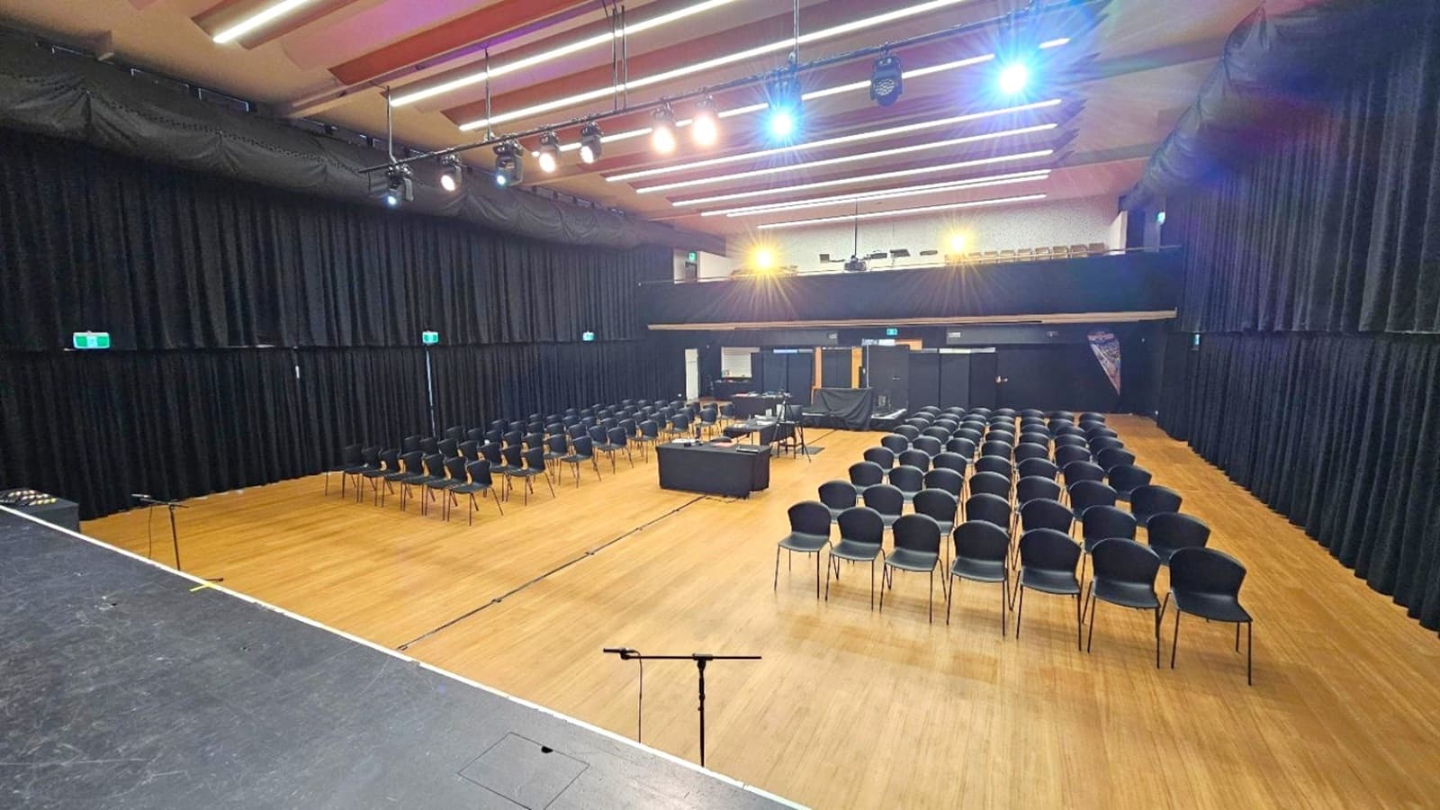 A large auditorium with a wooden floor and a high ceiling featuring rows of lights. On the left, there is a stage with a microphone stand. In the centre, rows of black chairs are arranged facing the stage, with a table in the middle. The background has a control area with equipment and a banner. Black curtains line the walls, and there are exit signs visible. The lighting is bright, illuminating the entire space.