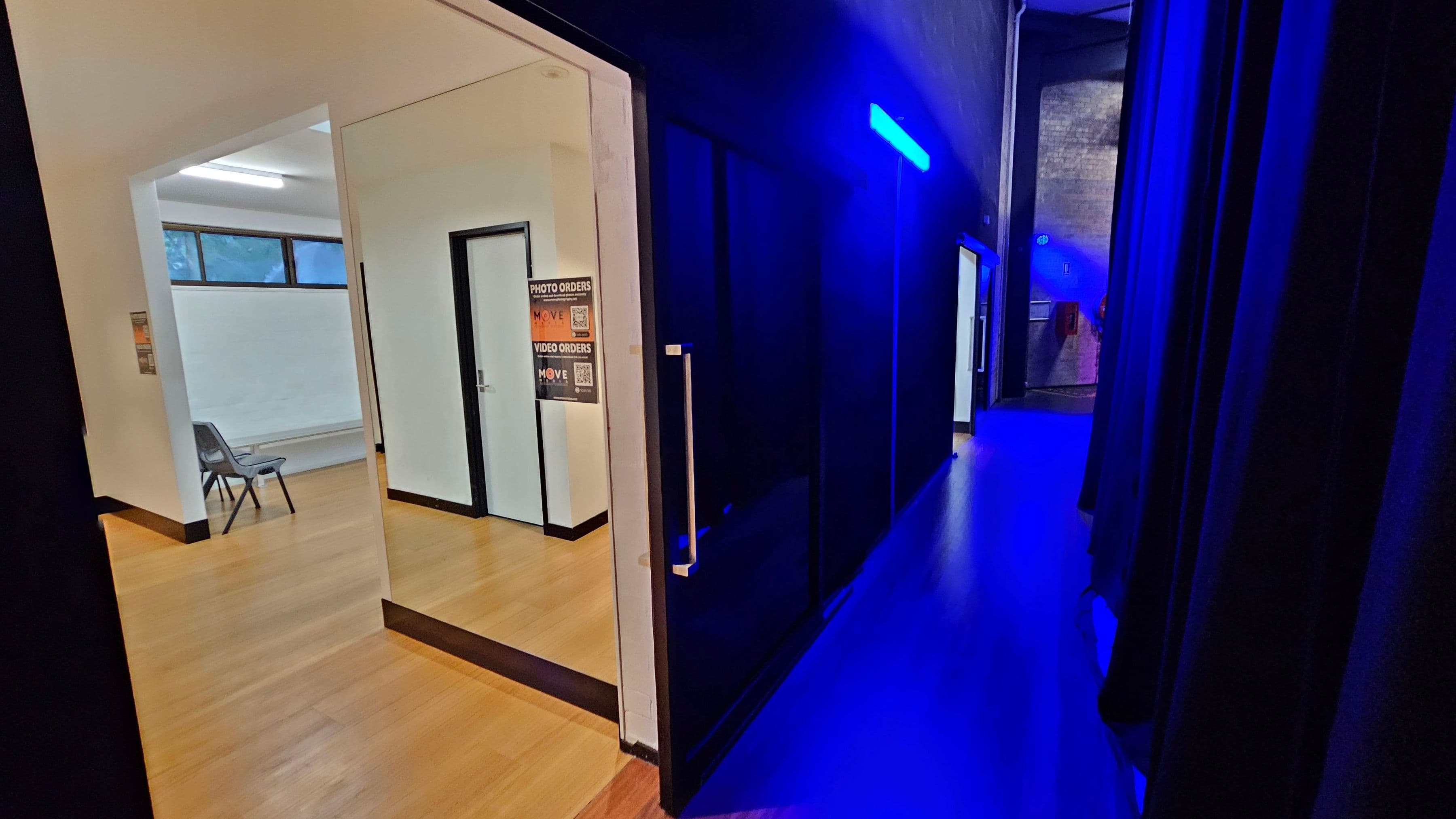 A hallway with a wooden floor and a black wall on the right, illuminated by blue lighting. On the left, there's an open doorway leading to a room with white walls, a fluorescent light, and a window with frosted glass. Inside the room, there are two grey chairs and a poster on the wall. The hallway extends into the distance, with more doors visible along the right side.