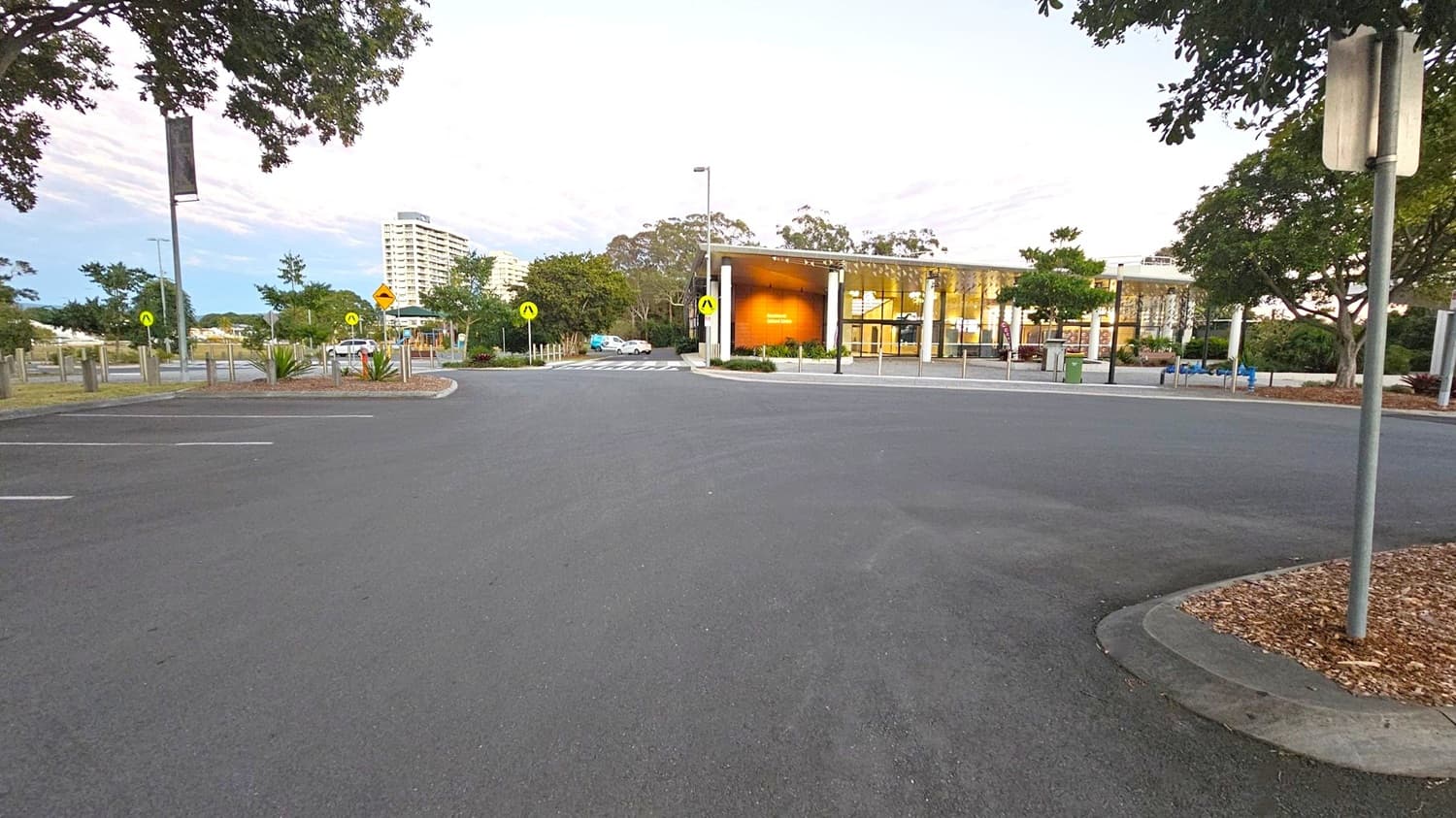 A wide, empty car park with a smooth asphalt surface. In the background, a modern building with large glass windows and a wooden facade is visible. To the left, there are trees and a few parked cars. On the right, more trees and a signpost are present. The area is well-lit with street lamps, and there are pedestrian crossing signs near the building. A tall building is visible in the distance.