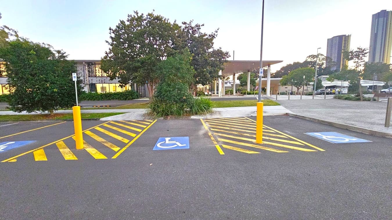 Three parking spaces marked for disabled access with blue wheelchair symbols on the ground. Yellow striped lines and yellow bollards separate the spaces. In the background, there are trees and a building with large windows. To the right, tall buildings are visible in the distance. The ground is asphalt, and the area is well-lit with streetlights.