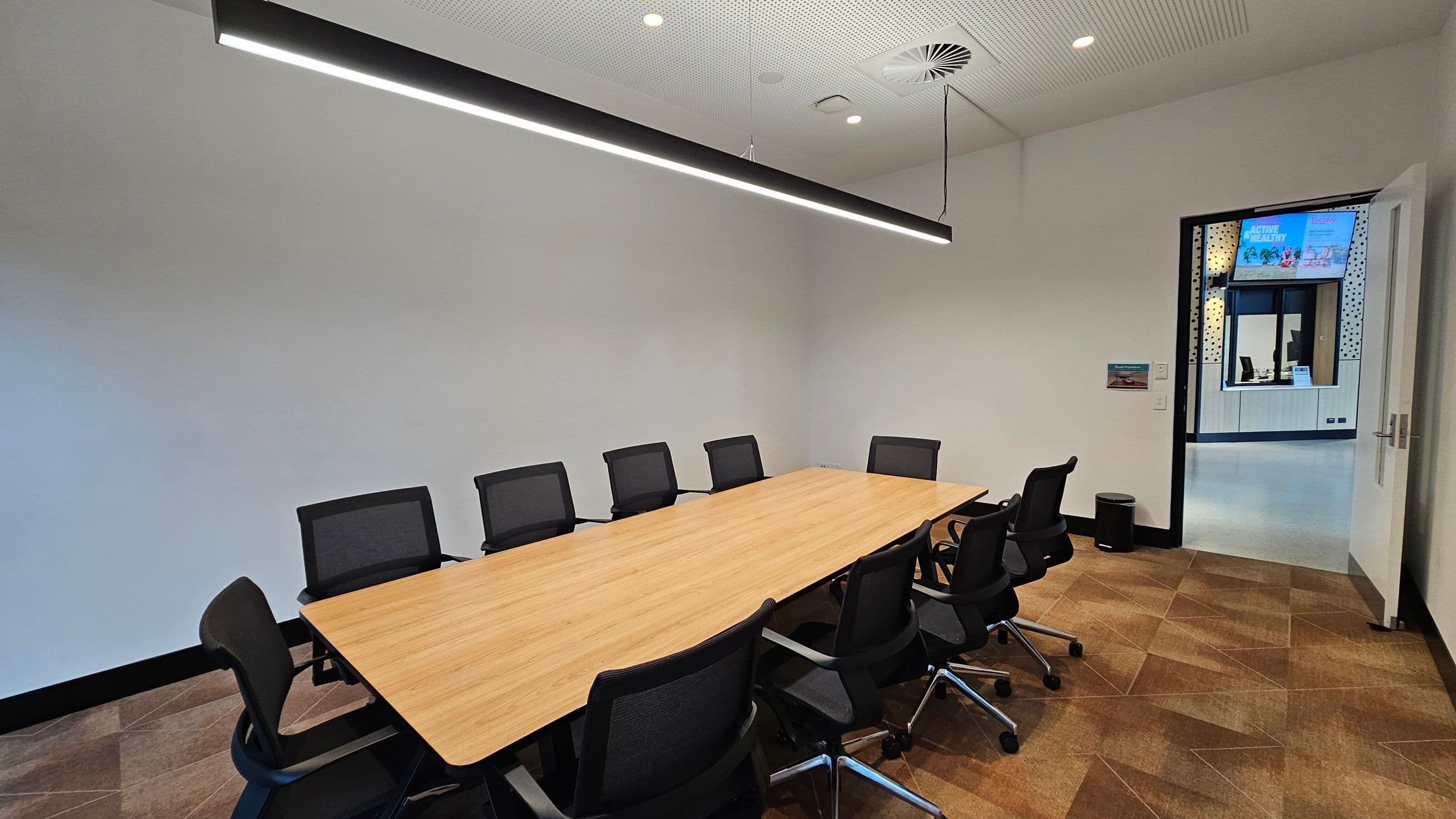 A modern conference room with a long wooden table surrounded by black office chairs on a patterned carpeted floor. A linear light fixture hangs from the ceiling, which is white with recessed lighting and a ventilation unit. An open door on the right leads to another room with visible signage and a window. A small waste bin is near the door. The walls are plain and light-coloured.