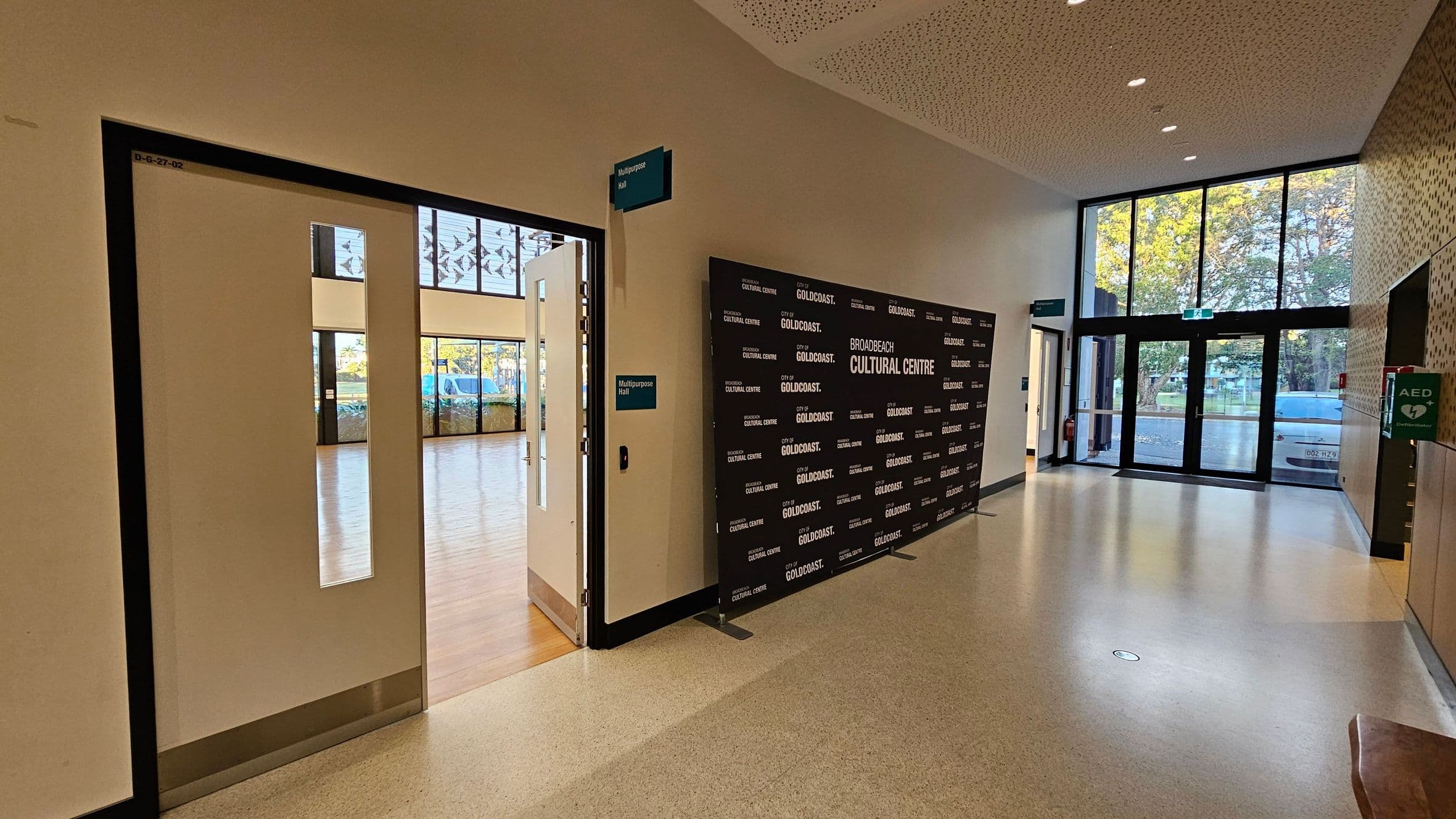 A hallway with a polished floor and a perforated ceiling. On the left, an open door leads to a room with wooden flooring and large windows. A sign above the door reads "Multipurpose Hall." In the centre, a black backdrop with "Broadbeach Cultural Centre" and "City of Gold Coast" logos. On the right, large glass doors open to the outside, with trees visible. An AED sign is mounted on the wall near the entrance.
