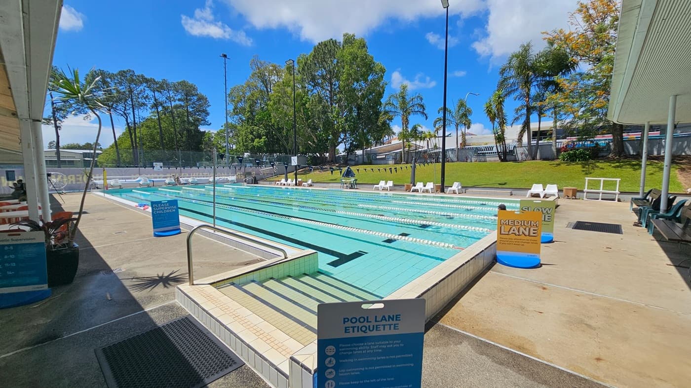 An outdoor swimming pool with multiple lanes, each marked by floating dividers. Signs indicate lane speeds, such as "Medium Lane." On the right, there are white lounge chairs and a grassy area with palm trees. On the left, a shaded area with seating and a potted plant. The pool deck is concrete, and there are signs about pool lane etiquette. The background features tall trees and a fence.