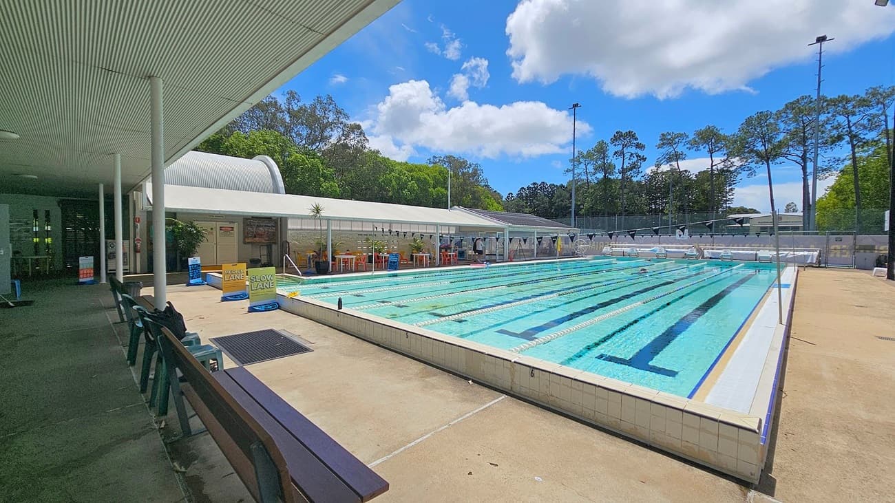 Outdoor swimming pool with multiple lanes, surrounded by a tiled deck. On the left, there's a covered area with benches and chairs. Signs indicate "Medium Lane" and "Slow Lane" near the pool's edge. In the background, a row of trees and a fence line the perimeter. The ceiling of the covered area is corrugated, and the floor is concrete. Bright sunlight casts shadows across the scene.