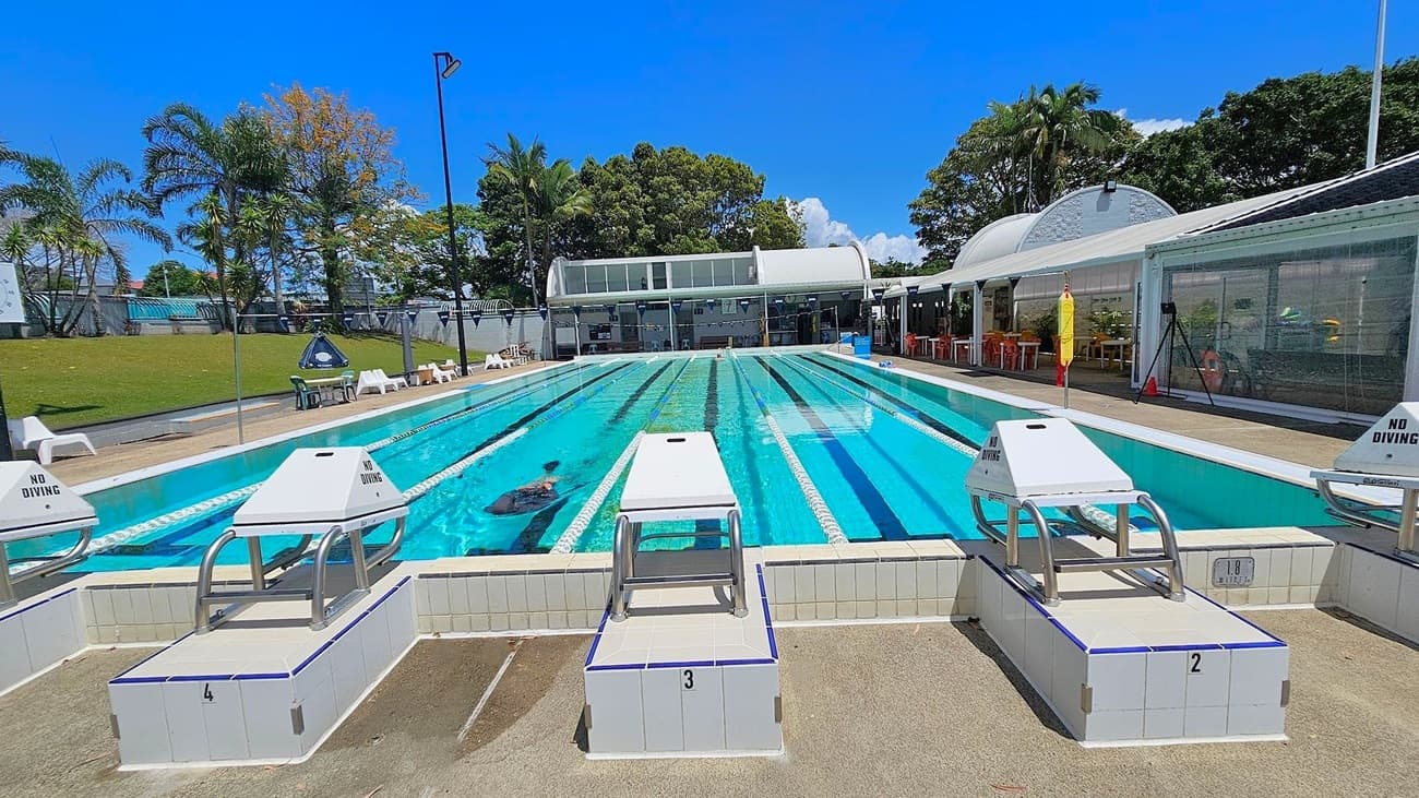 An outdoor swimming pool with multiple lanes, each marked by lane ropes. In the foreground, there are starting blocks numbered 2, 3, and 4, with "No Diving" signs. A swimmer is visible in the water. The background features a building with large windows and a curved roof, surrounded by trees and greenery. White chairs and a lifeguard stand are positioned along the poolside. The area is well-lit, suggesting a sunny day.