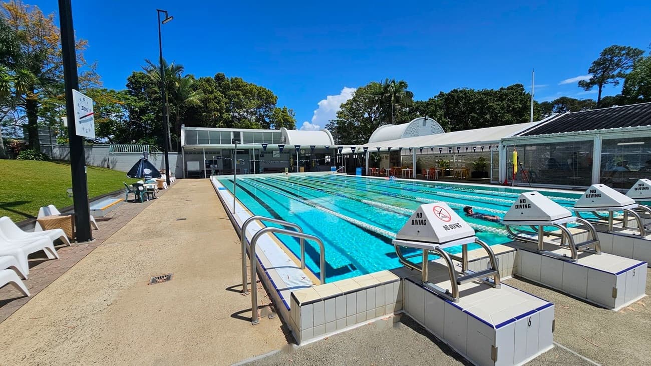 Outdoor swimming pool with clear blue water and marked lanes. On the right, there are starting blocks with "No Diving" signs. A swimmer is in one of the lanes. The left side has a paved area with white lounge chairs and a few tables with chairs. In the background, there are trees and a building with a curved roof. The pool area is surrounded by greenery.