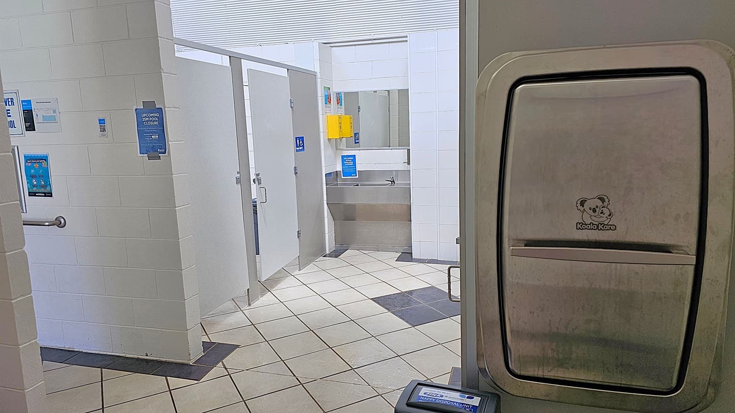 A narrow corridor with white tiled walls and metal handrails on both sides. At the end, there is a wall with a sign reading "PLEASE SHOWER BEFORE ENTERING POOL" and several posters. A soap dispenser is mounted on the left wall above a stainless steel sink. Below the sink is a white bin with a black liner. The floor has a black and white diamond pattern. On the right, a turquoise metal gate with a padlock is visible.