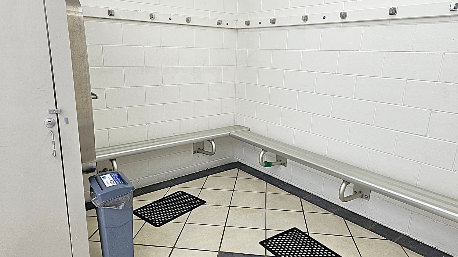 A changing room with white tiled walls and a corner bench made of metal. The bench is supported by metal brackets. Above the bench, there are hooks for hanging items. The floor is tiled with a light colour and has two black rubber mats. On the left, a door is partially open, and a small bin with a liner is positioned near the entrance.