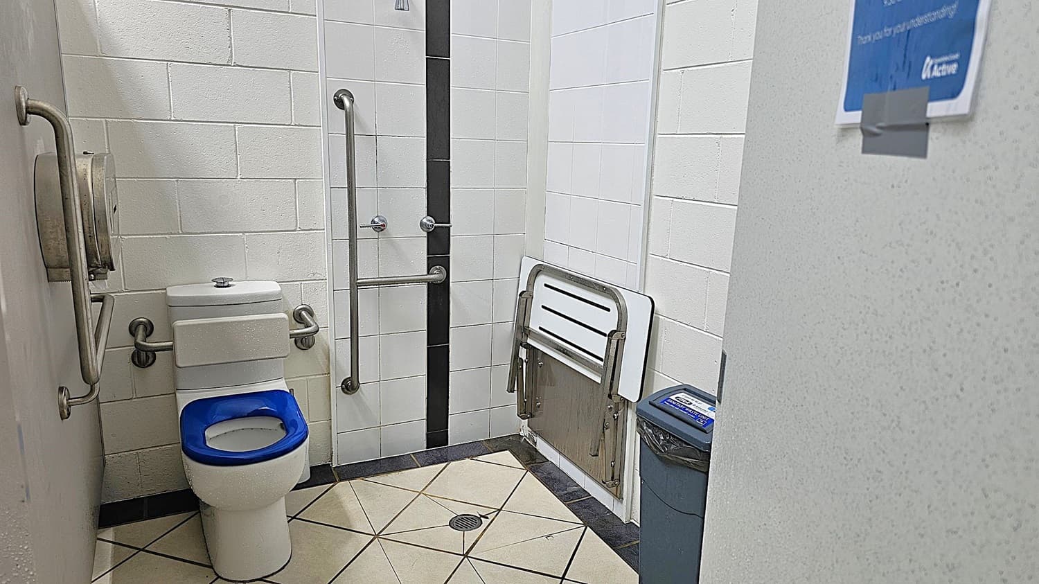 A public restroom with white tiled walls and a tiled floor. On the left, there is a toilet with a blue seat and metal grab bars on both sides. A toilet paper dispenser is mounted on the wall. In the centre, a vertical black tile strip runs down the wall. On the right, a foldable baby changing table is attached to the wall above a bin. A partial view of a door with a blue sign is visible on the far right.