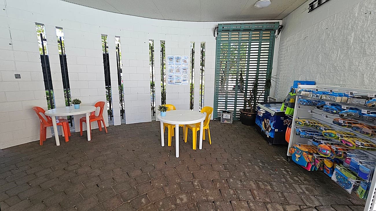 A small shop interior with a metal counter in the foreground. On the left, there are sunscreen products and brochures. Behind the counter, a fridge stocked with drinks is visible, along with a coffee machine and a small fridge. On the right, a snack display with crisps and sweets is next to a first aid sign. The background shows an open window with a view of greenery outside. The ceiling is lined with fluorescent lights.
