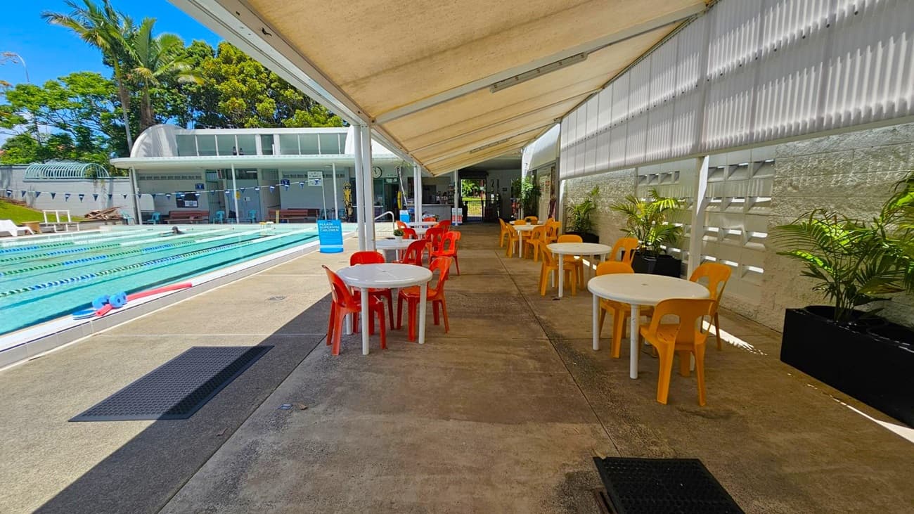 A swimming pool area with lanes on the left, surrounded by greenery and a building with a curved roof. On the right, there is a covered seating area with white round tables and colourful plastic chairs, mostly orange and yellow. Potted plants line the wall, which has a textured design. The floor is concrete, and the ceiling is a white canopy providing shade.