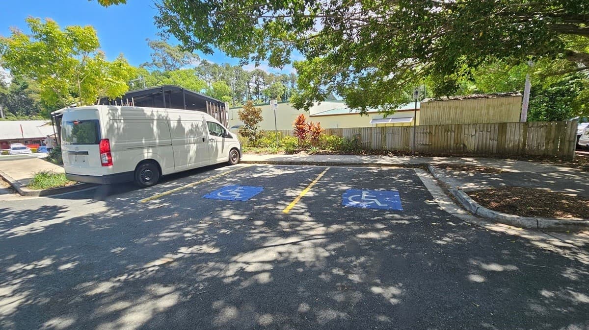 A white van is parked on the left in a parking area with two marked disabled parking spaces in the centre. The ground is asphalt with blue disabled symbols. In the background, there is a wooden fence and some greenery, including trees and bushes. Shadows from the trees are cast on the ground, and a building with a light green wall is visible behind the fence.